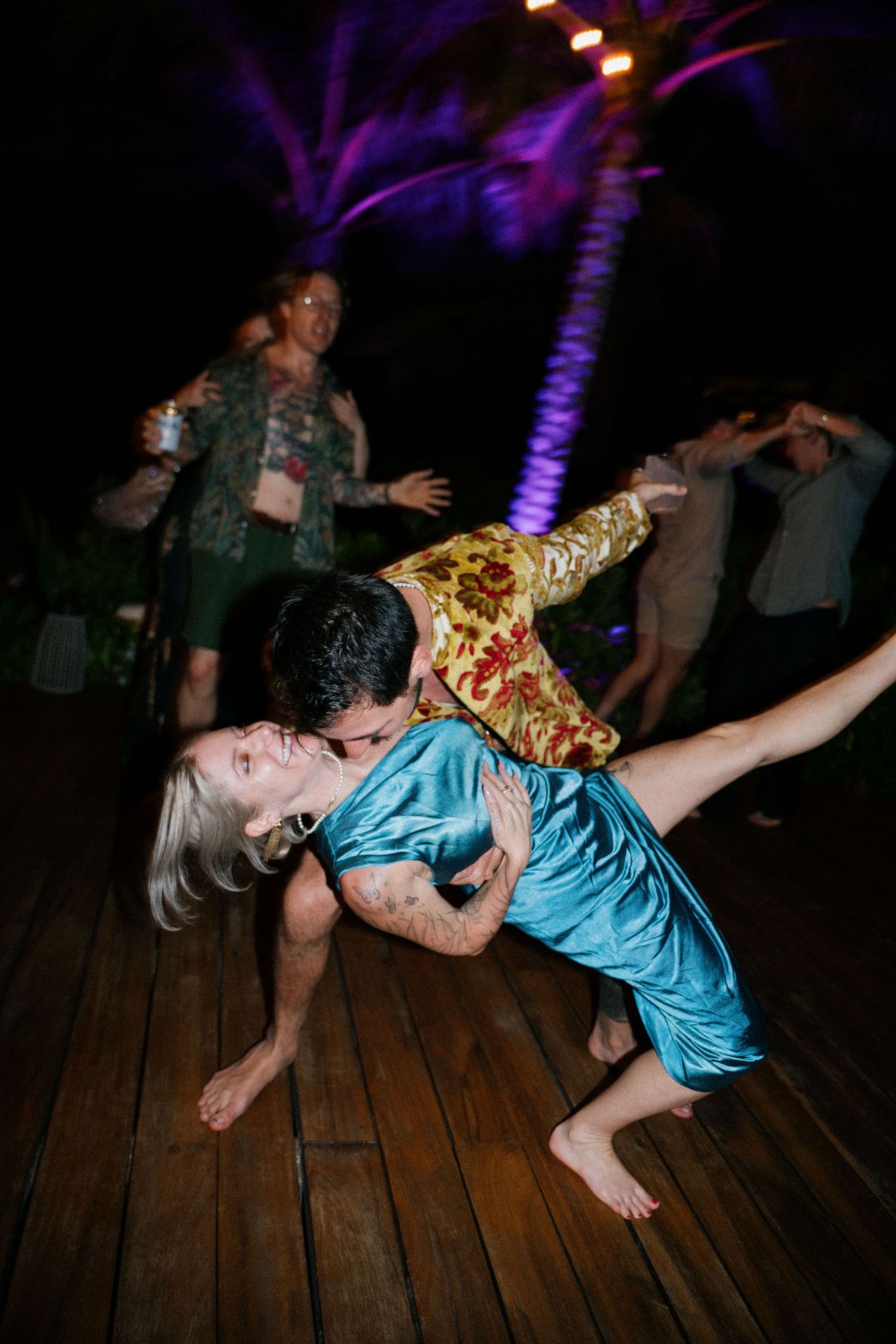 Dancing couple performs dramatic dip move on wooden deck under lit palm trees at night.