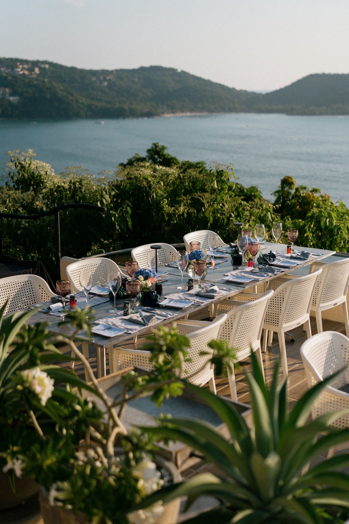 Elegant outdoor dining table setup overlooking a lake with mountain views and tropical plants.