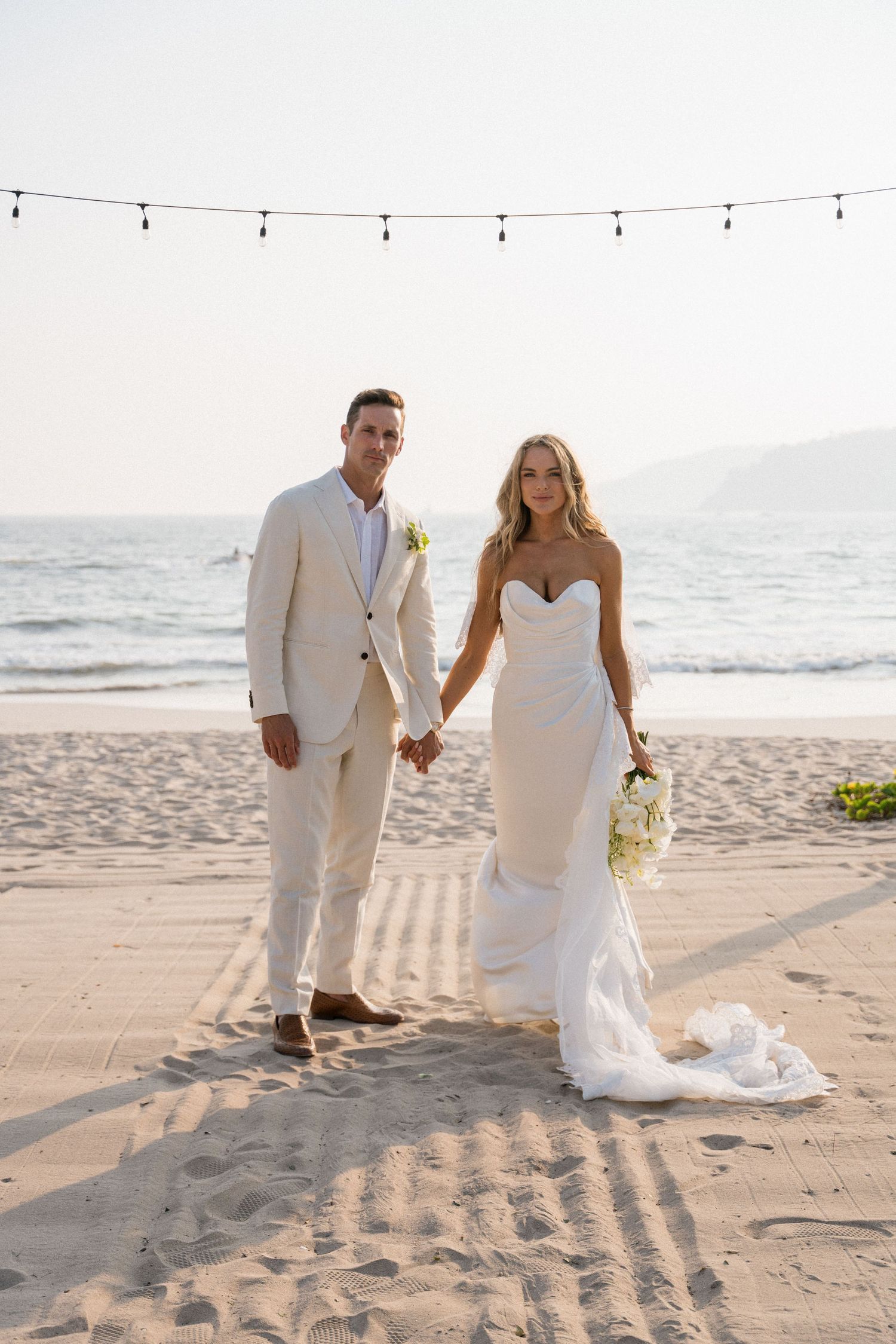 A couple in wedding attire walking on a sandy beach at sunset.