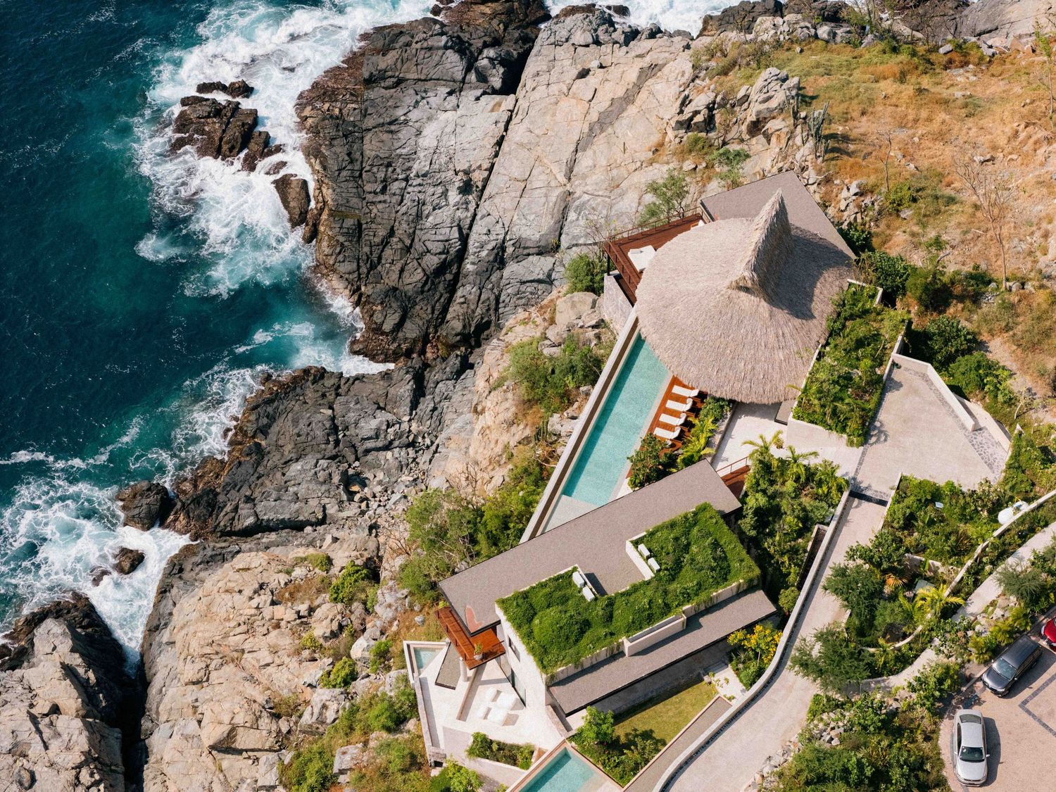 Aerial view of a luxury cliffside villa with infinity pool overlooking dramatic rocky coastline and turquoise ocean waters.
