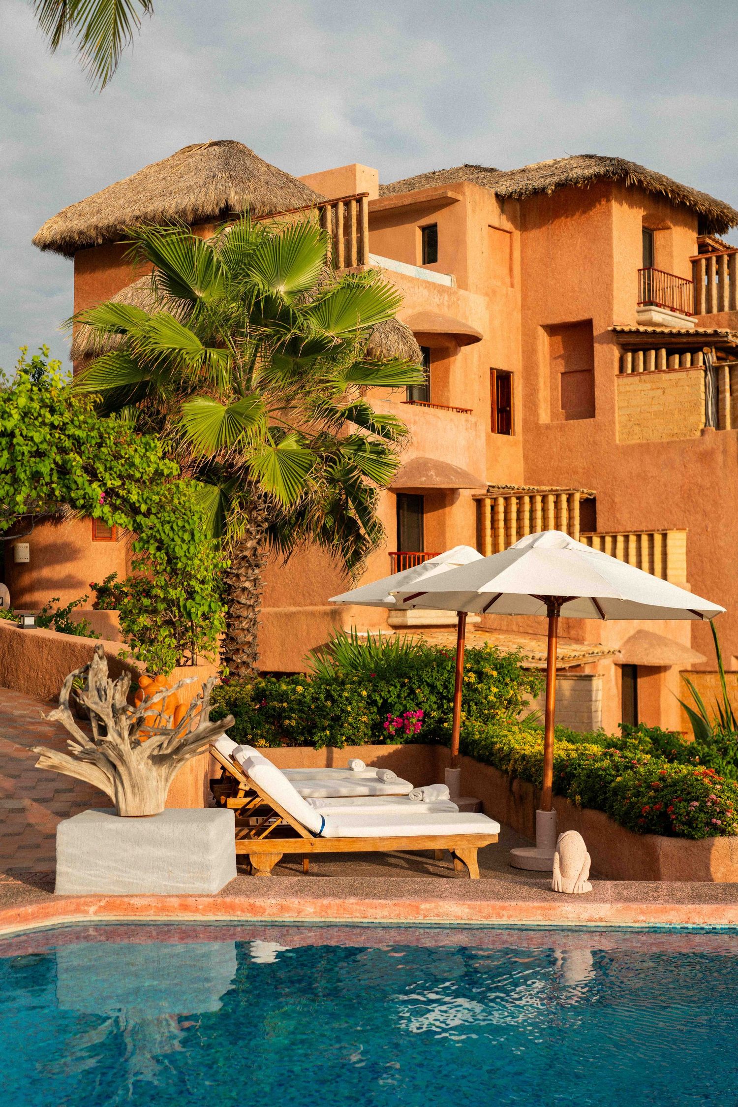 Luxurious terracotta resort villa with thatched roof and poolside lounge chairs under white umbrella by palm trees.