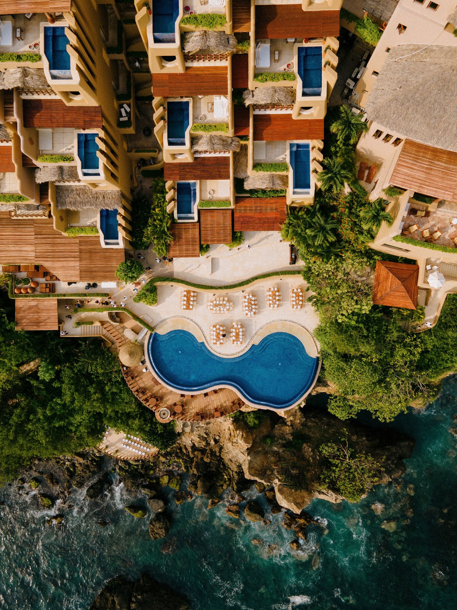 Aerial view of luxury oceanfront resort with curved swimming pool and terracotta buildings.