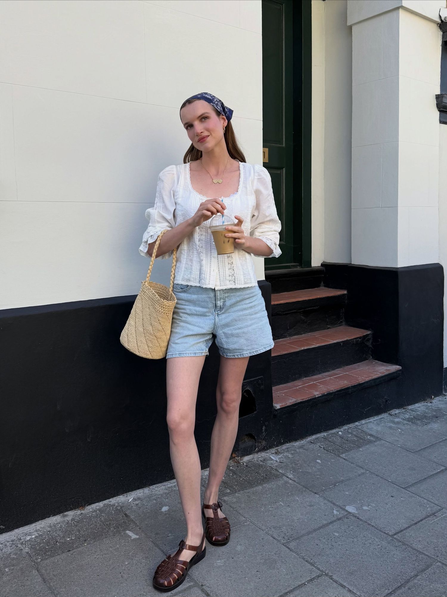 Woman in white top, denim shorts, brown sandals, and headscarf holding iced coffee with woven bag.