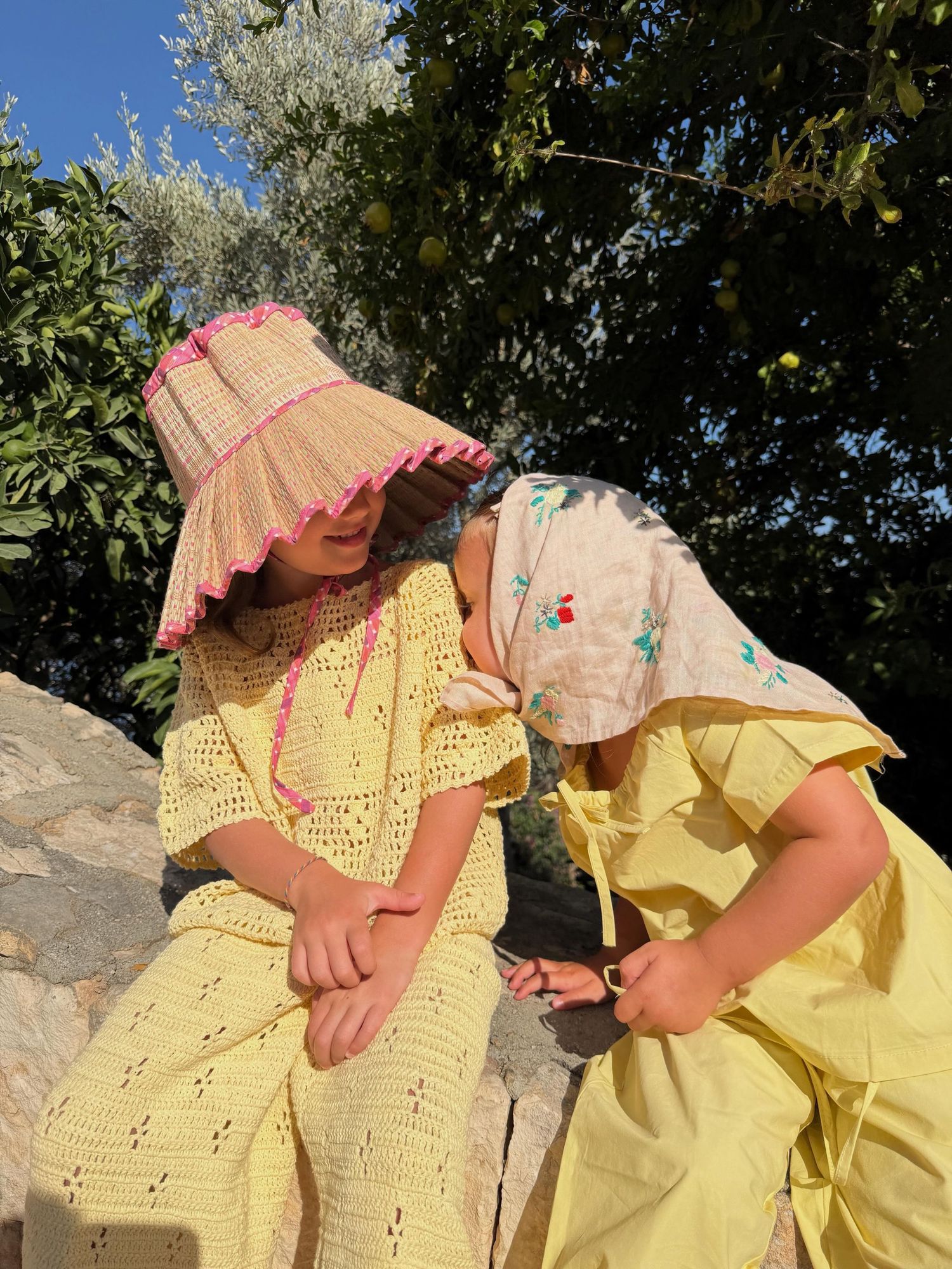 Two women in yellow outfits sit outdoors, one wearing a straw hat with pink trim, surrounded by lush green trees.