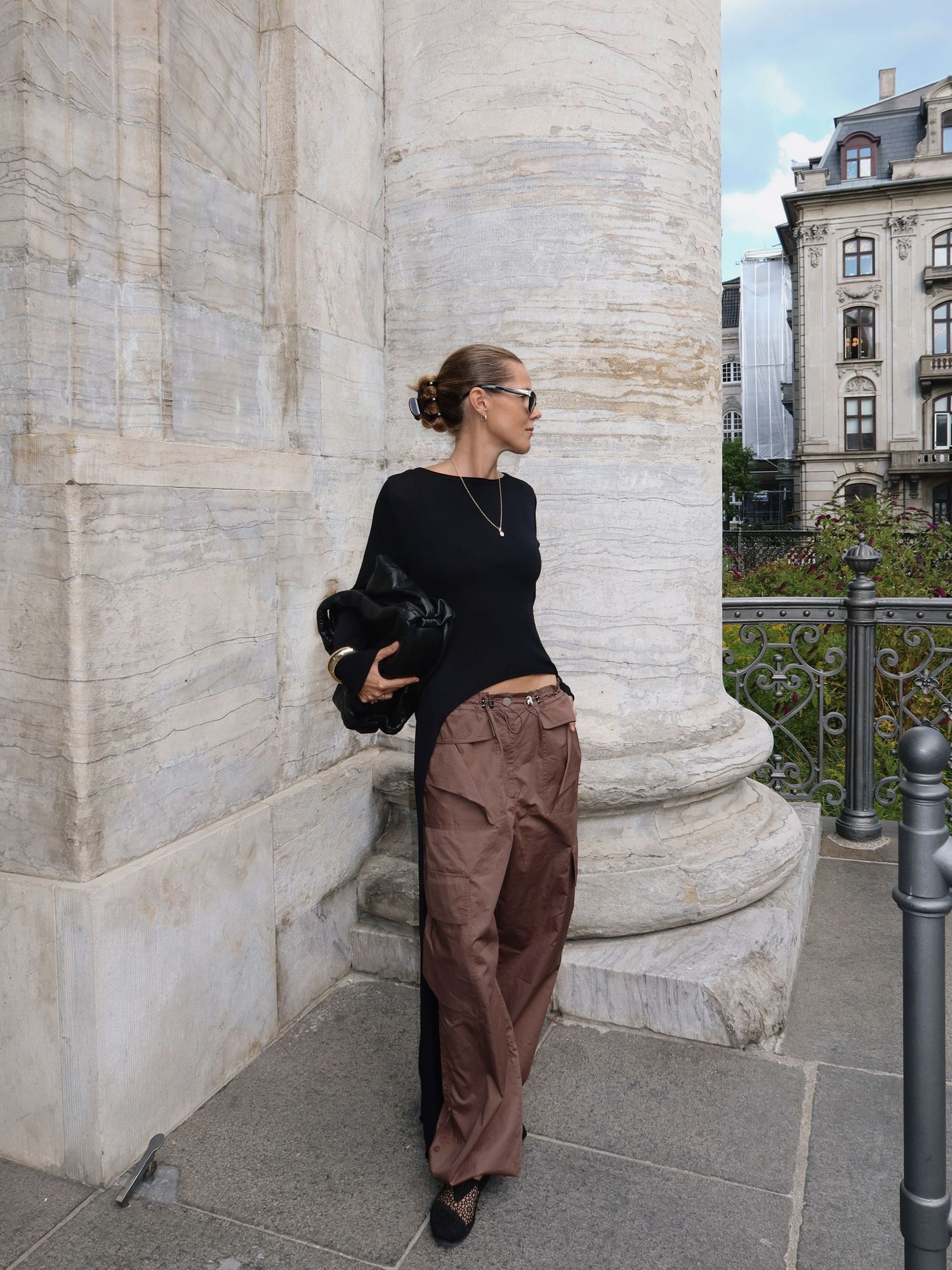 Woman in black top and brown cargo pants leaning against a stone column outdoors.