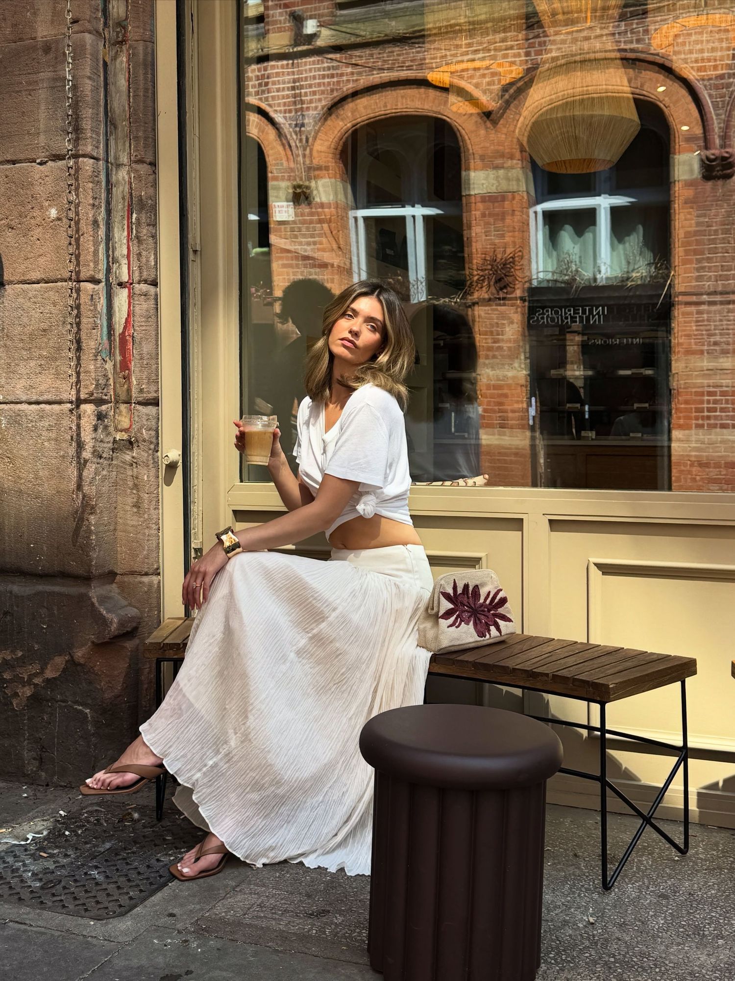 Woman in white crop top and maxi skirt sitting outside a cafe holding an iced coffee on a sunny day.
