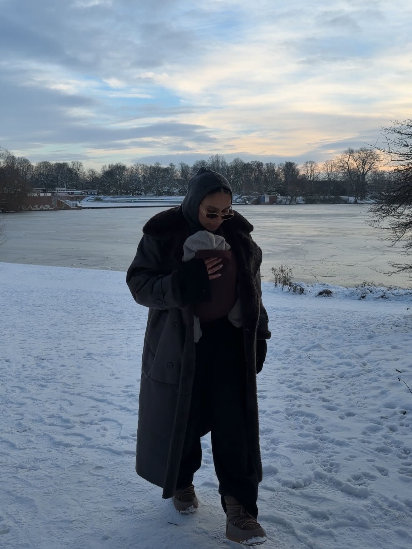 Person in long dark coat holding a drink, standing in snow near a frozen lake on a cloudy winter day.