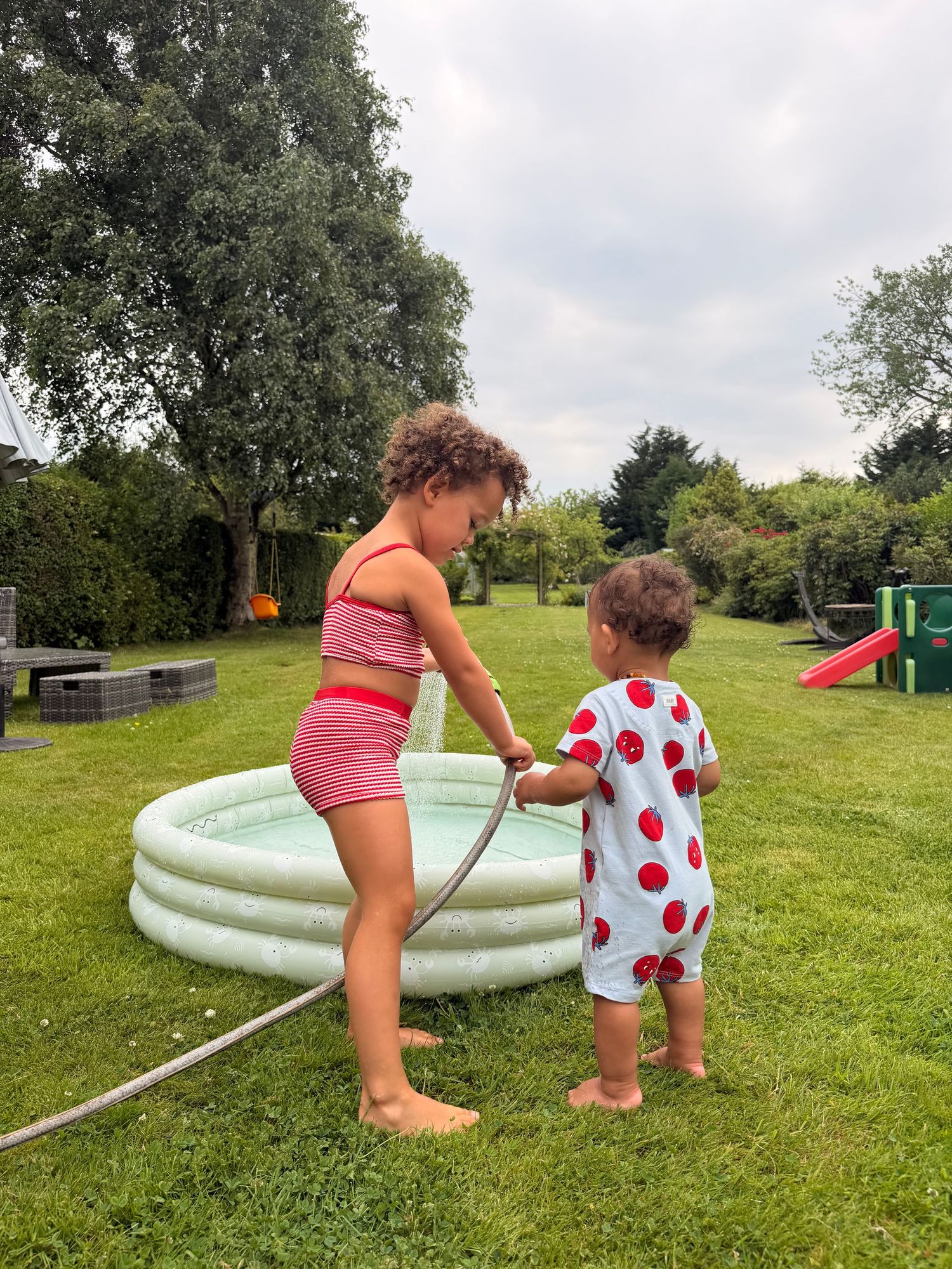 Girl in red swimsuit fills inflatable pool with hose while toddler in strawberry romper helps in sunny backyard.