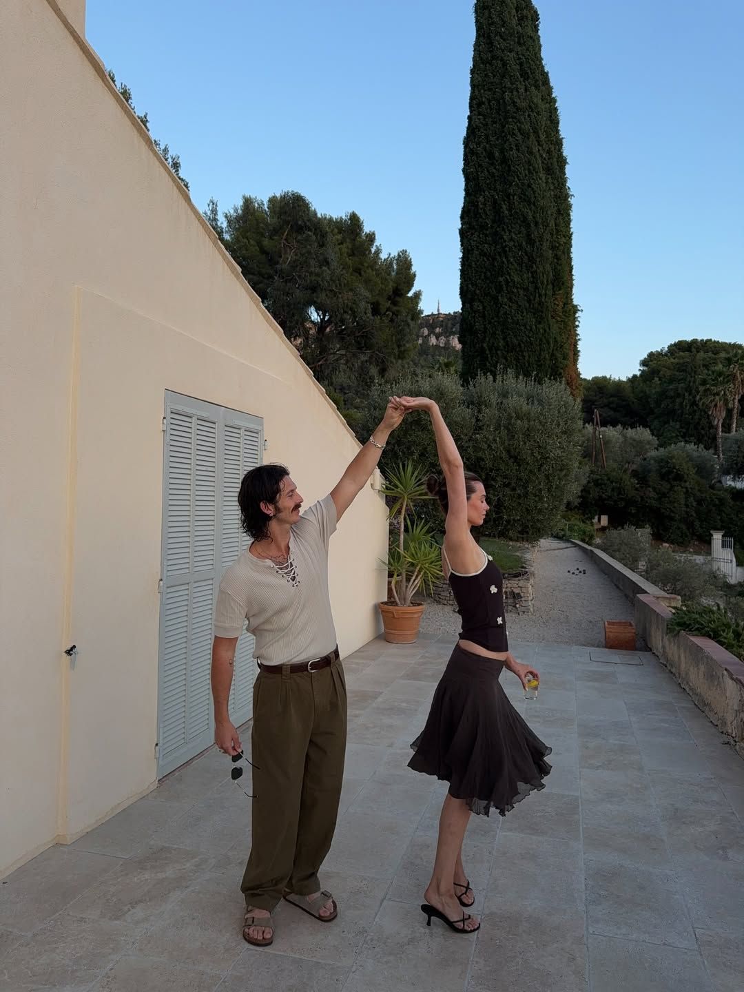 Couple dancing outdoors on a terrace with cypress trees and blue sky in Mediterranean setting.