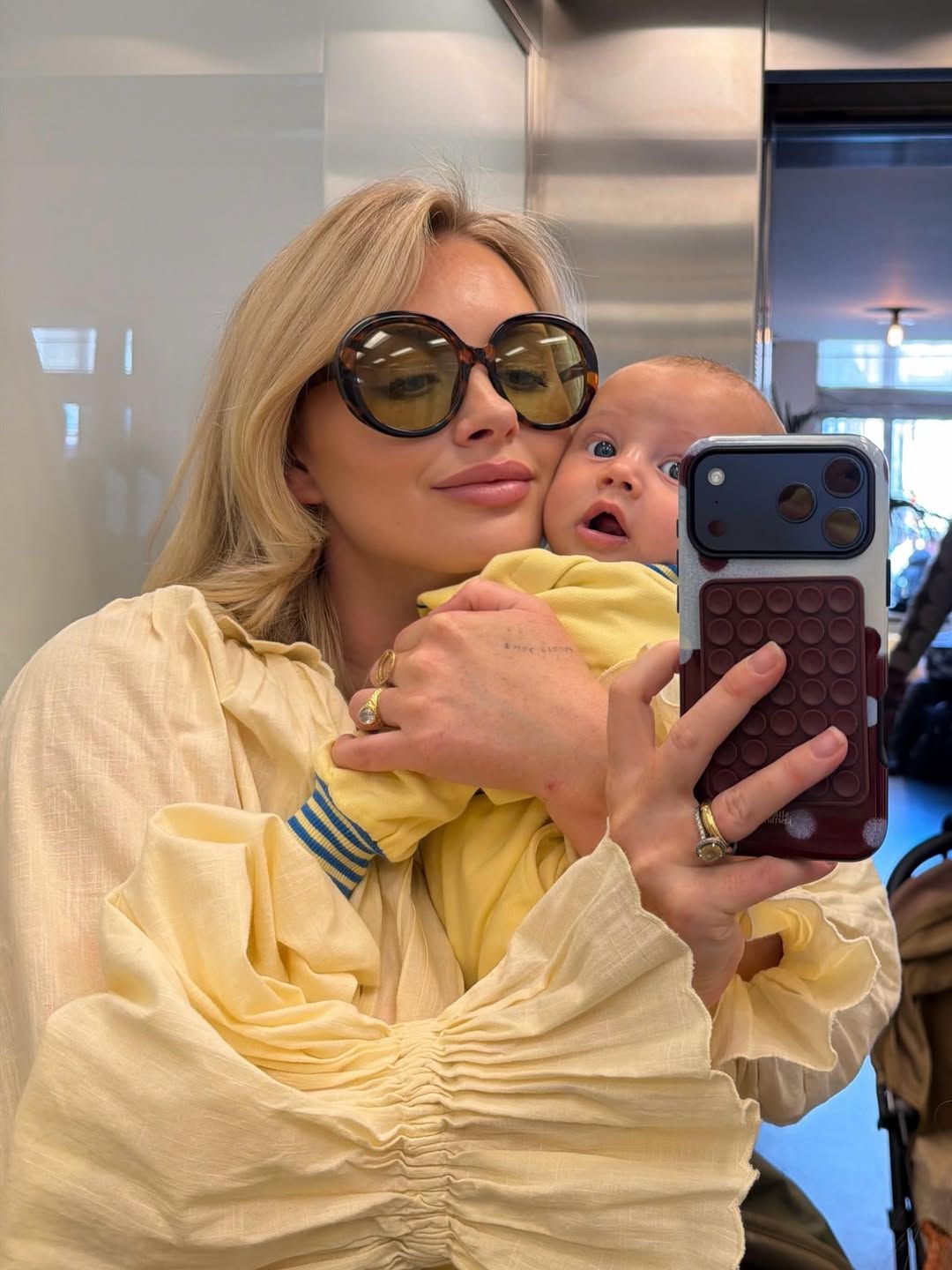 Blonde woman in large sunglasses holding a baby while taking a mirror selfie indoors.
