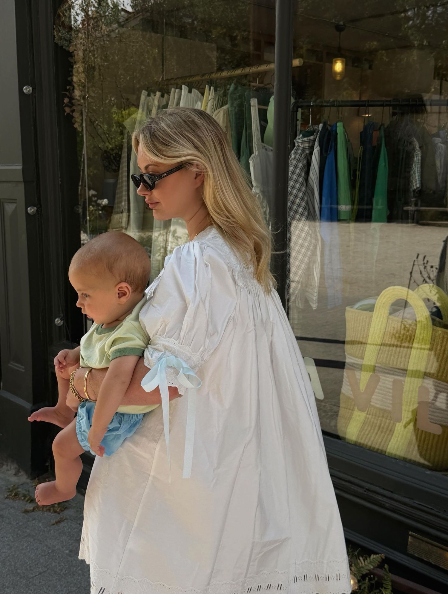 Blonde woman in white dress holding a baby outside a boutique shop window.