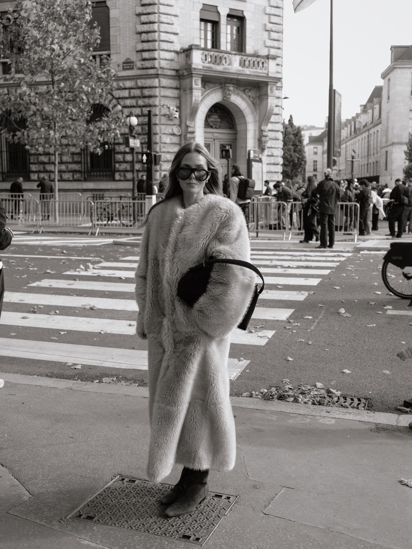 Woman in oversized faux fur coat and sunglasses posing on city street corner in black and white photo.