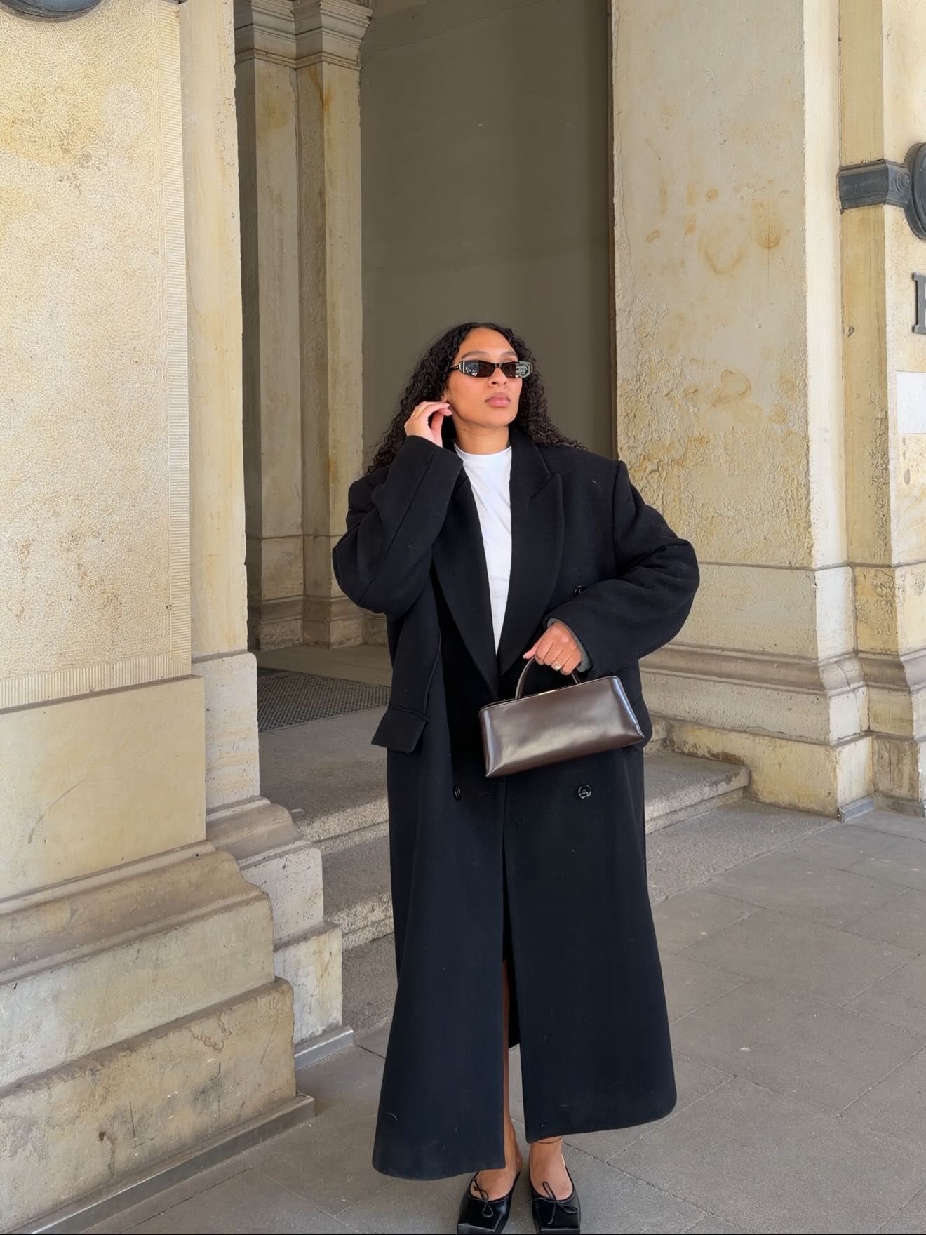 Woman in long black coat, white top, sunglasses, holding black leather bag, standing near stone columns.