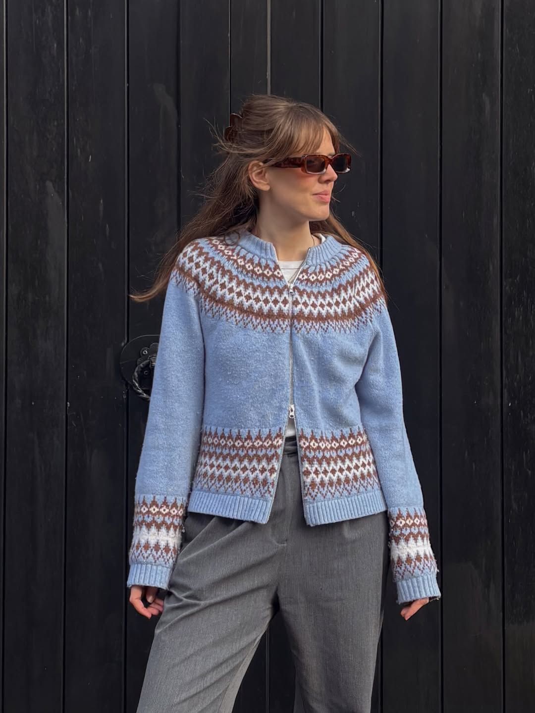 Woman wearing a light blue Fair Isle knit cardigan with brown pattern detail, paired with grey trousers and red sunglasses.