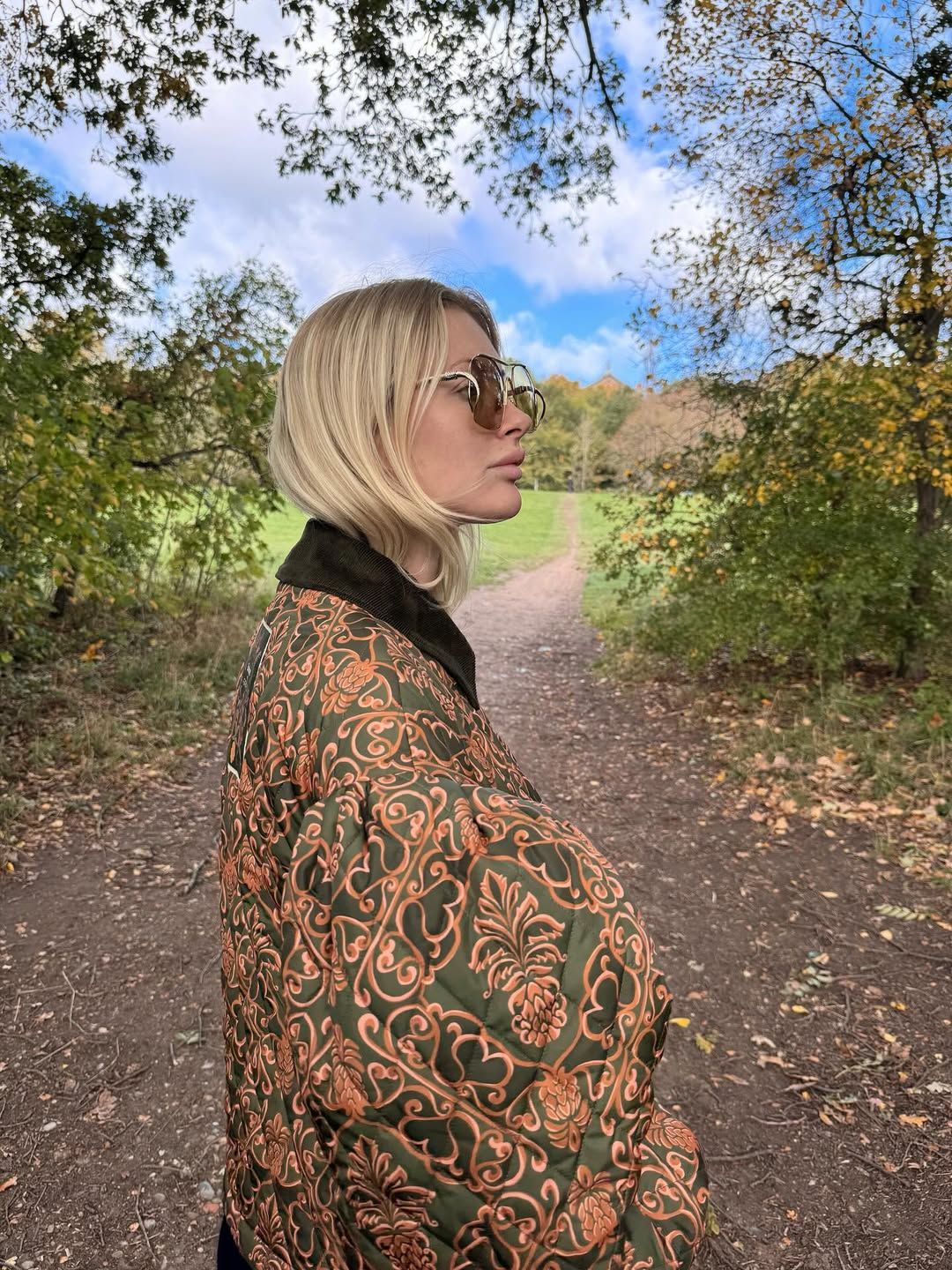 Blonde woman in patterned green jacket with glasses on autumn forest path, looking away.