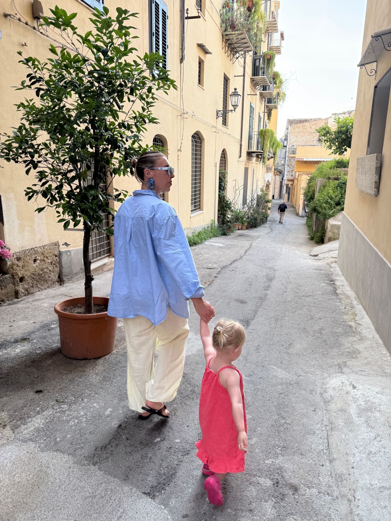 Adult and young child walk hand-in-hand down a charming narrow Italian alleyway with potted trees and yellow buildings.