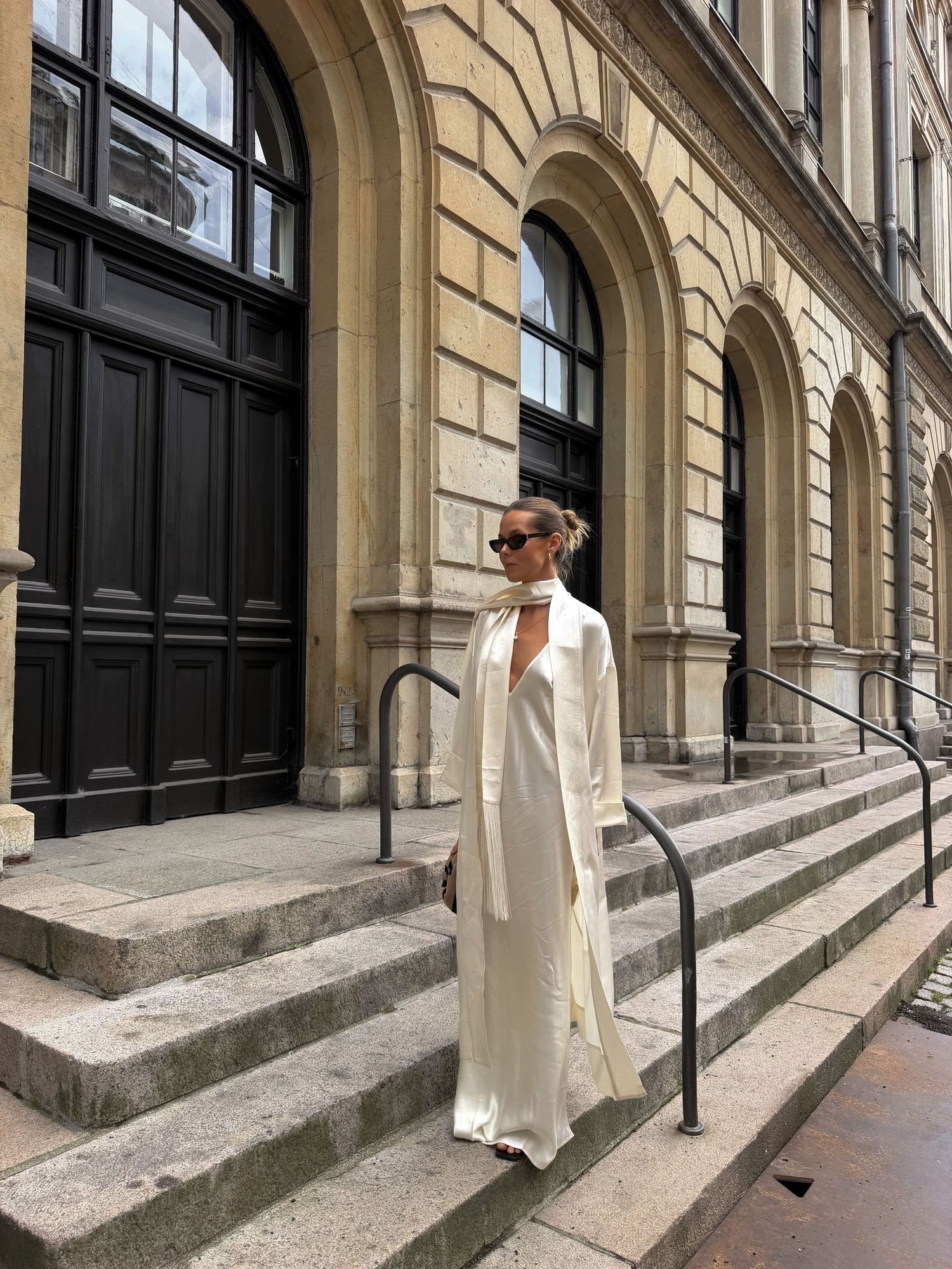Woman in white maxi dress and long coat standing on stone steps of a classic building.