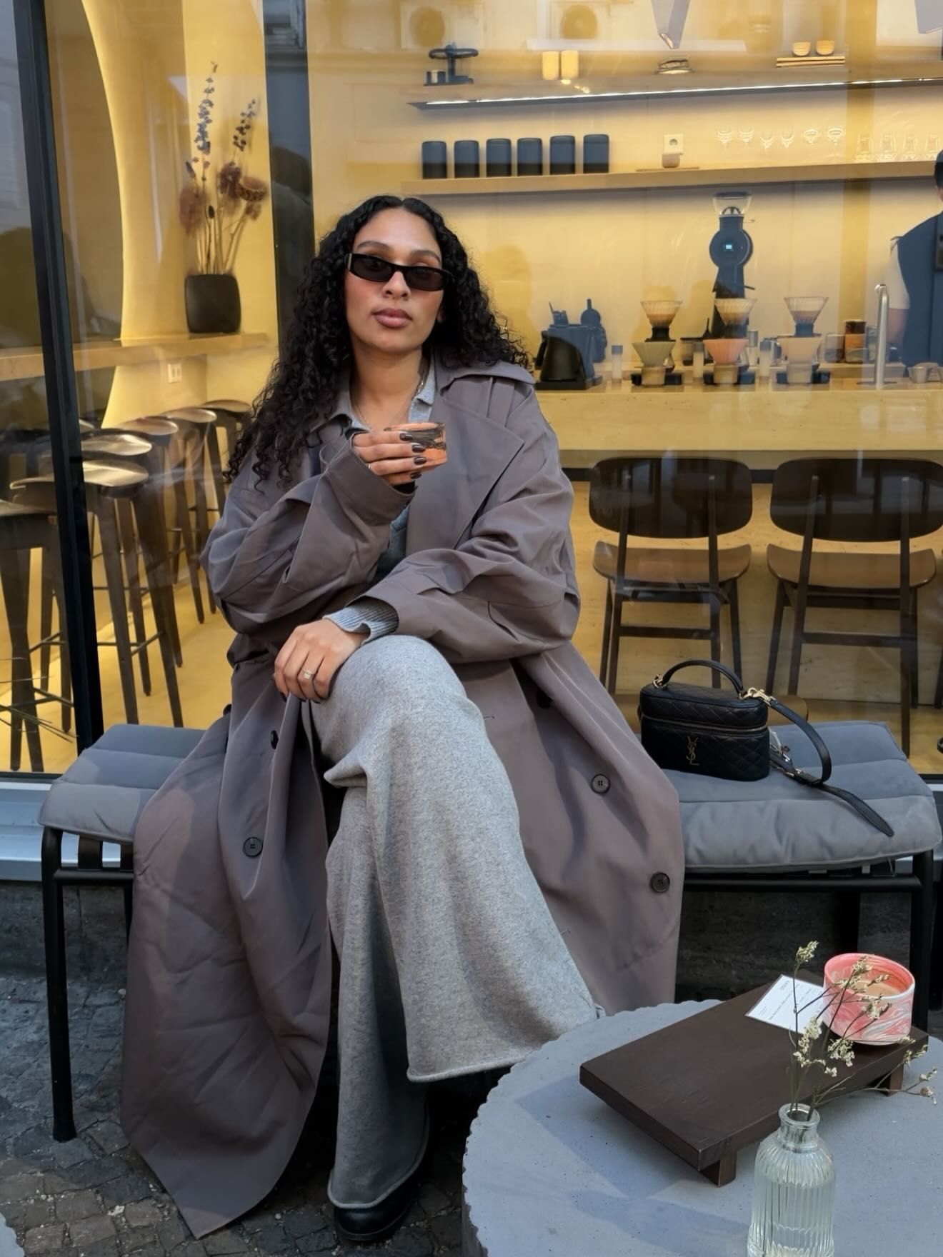 Woman in gray trench coat and sunglasses sitting at outdoor cafe table with black handbag.