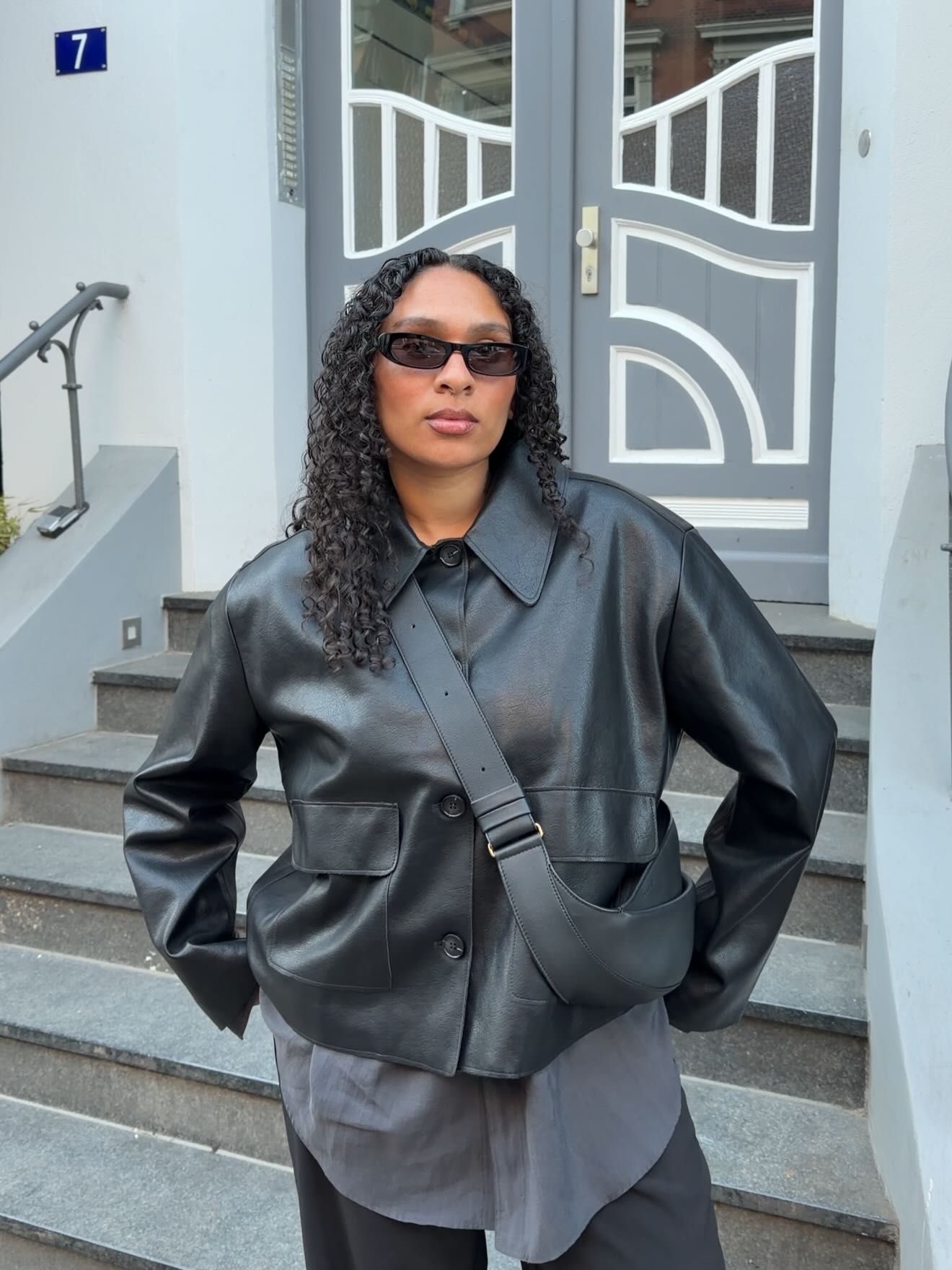 Woman in black leather jacket and crossbody bag poses on steps outside building with decorative door.