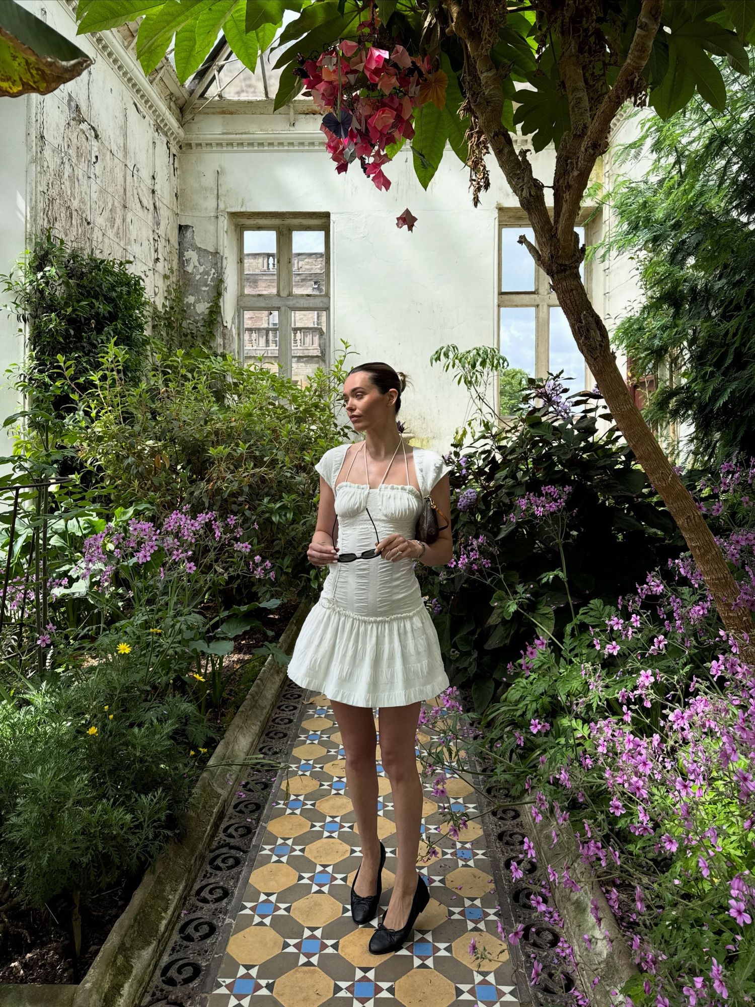 Woman in white mini dress standing on mosaic tile path surrounded by lush greenery and purple flowers.