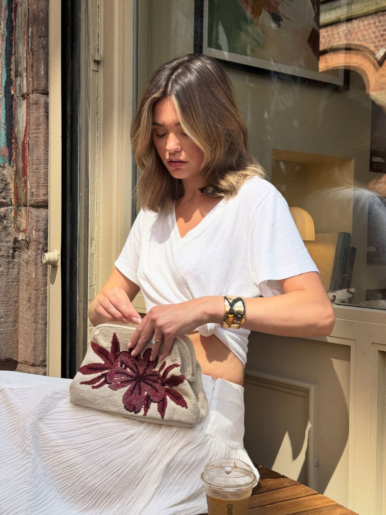 Woman in white top holding a beige clutch bag with purple floral embroidery at an outdoor cafe.