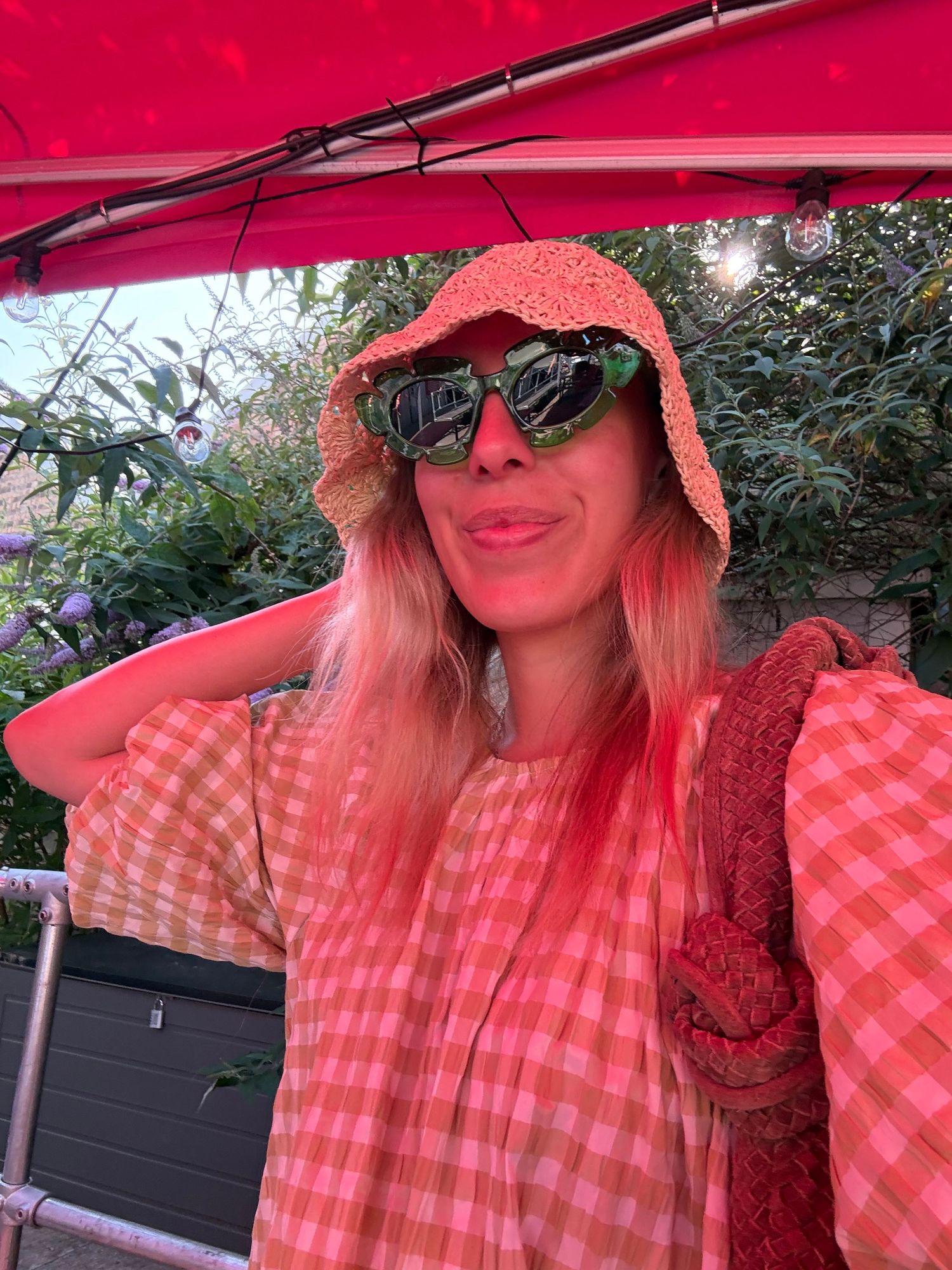 Woman in pink gingham dress and orange bucket hat with heart-shaped sunglasses posing under a red canopy outdoors.