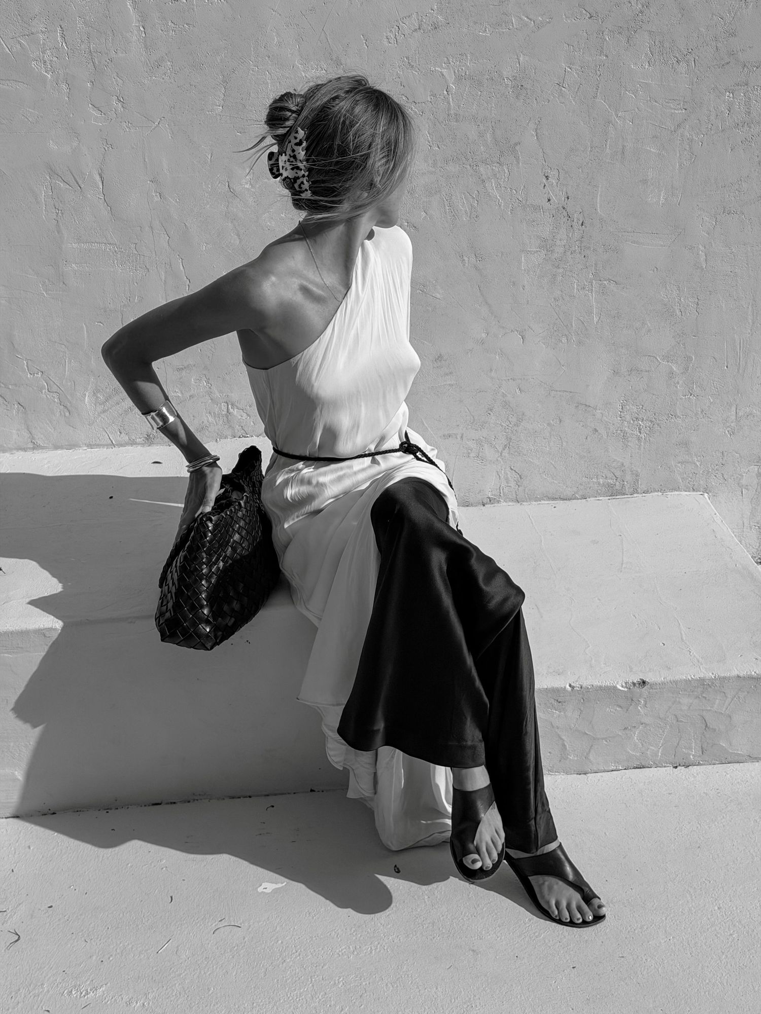 Woman in one-shoulder white top and wide-leg pants sits on white ledge holding woven bag, black and white photo.