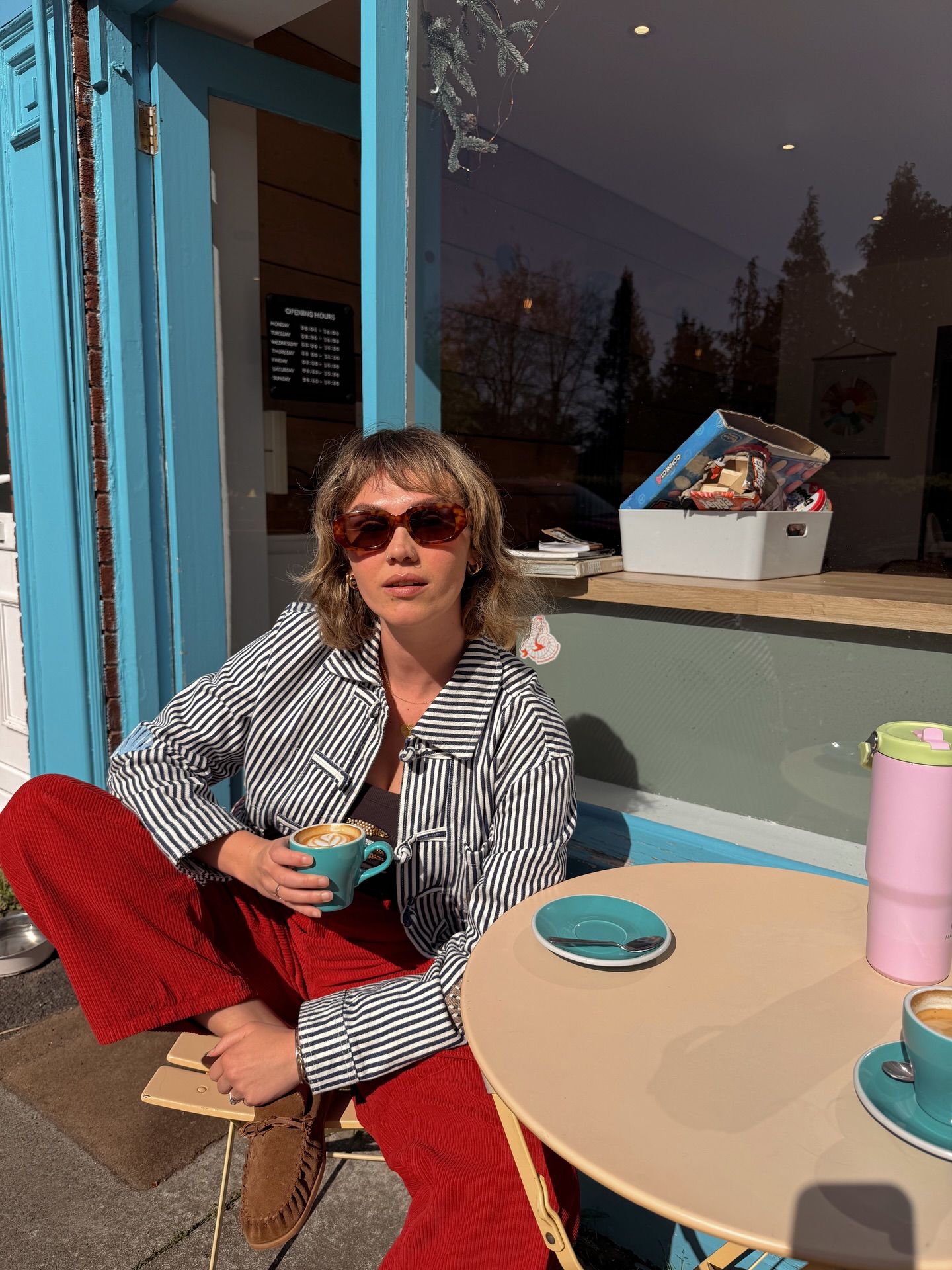 Woman in striped jacket and red pants sipping coffee at outdoor cafe table, wearing red sunglasses on sunny day.