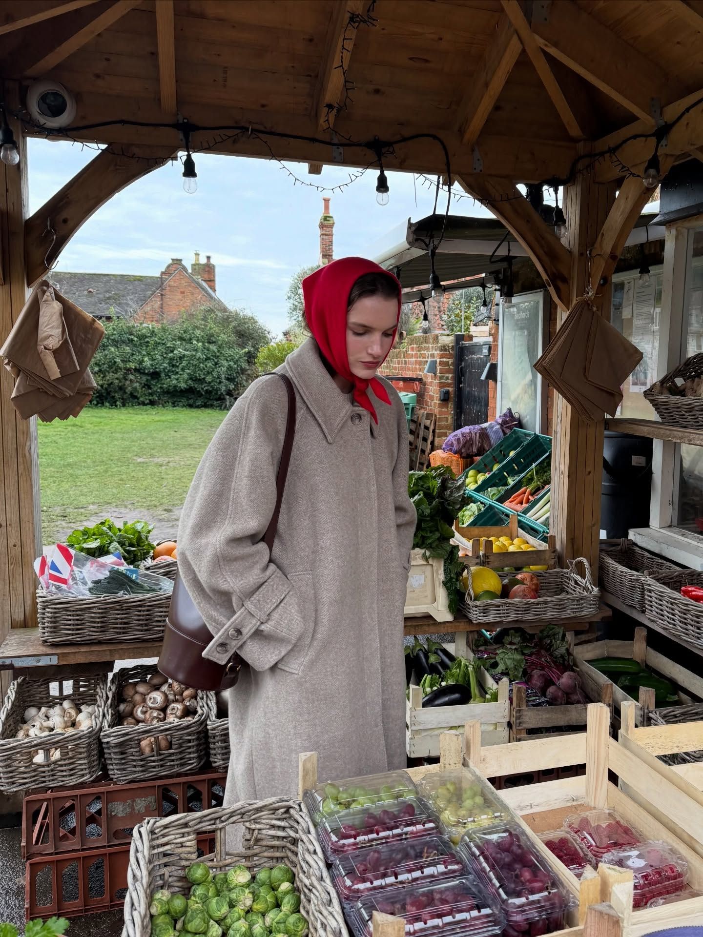 Person in gray coat and red headscarf browsing fresh vegetables at an outdoor farm stand market stall.
