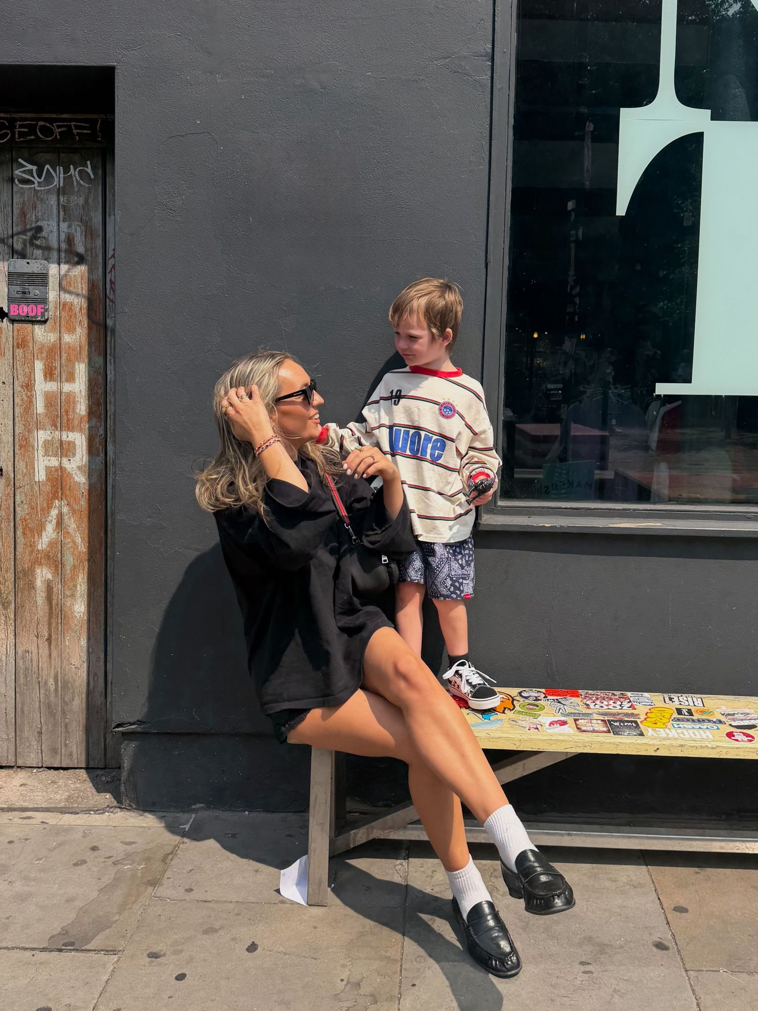 Woman in black dress and sunglasses sits on bench with young boy in striped sweater outside urban storefront.