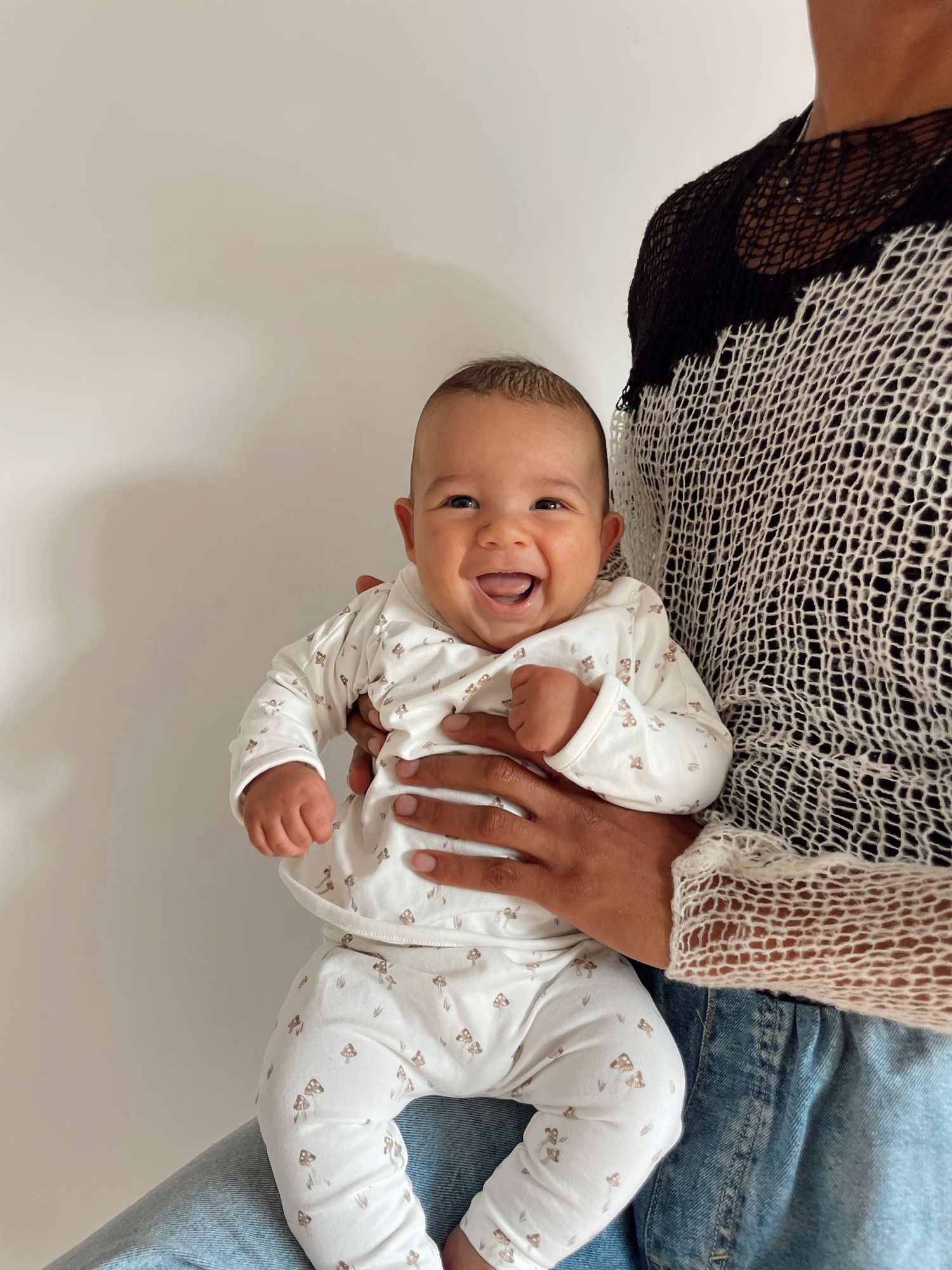 A baby in white floral pajamas smiles joyfully while being held against a white wall.
