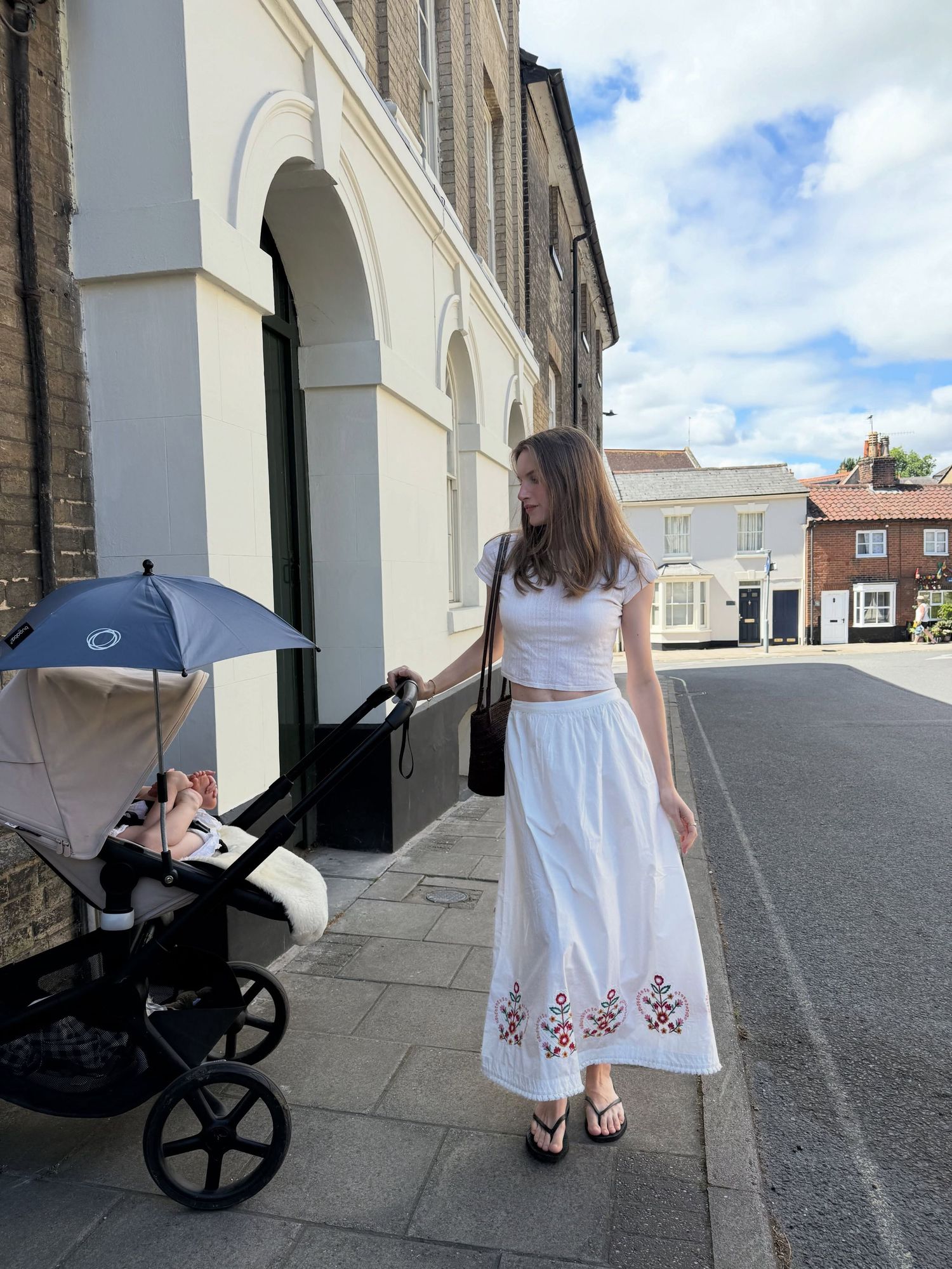 Woman in white crop top and embroidered midi skirt pushing a pram on a sunny cobblestone street.