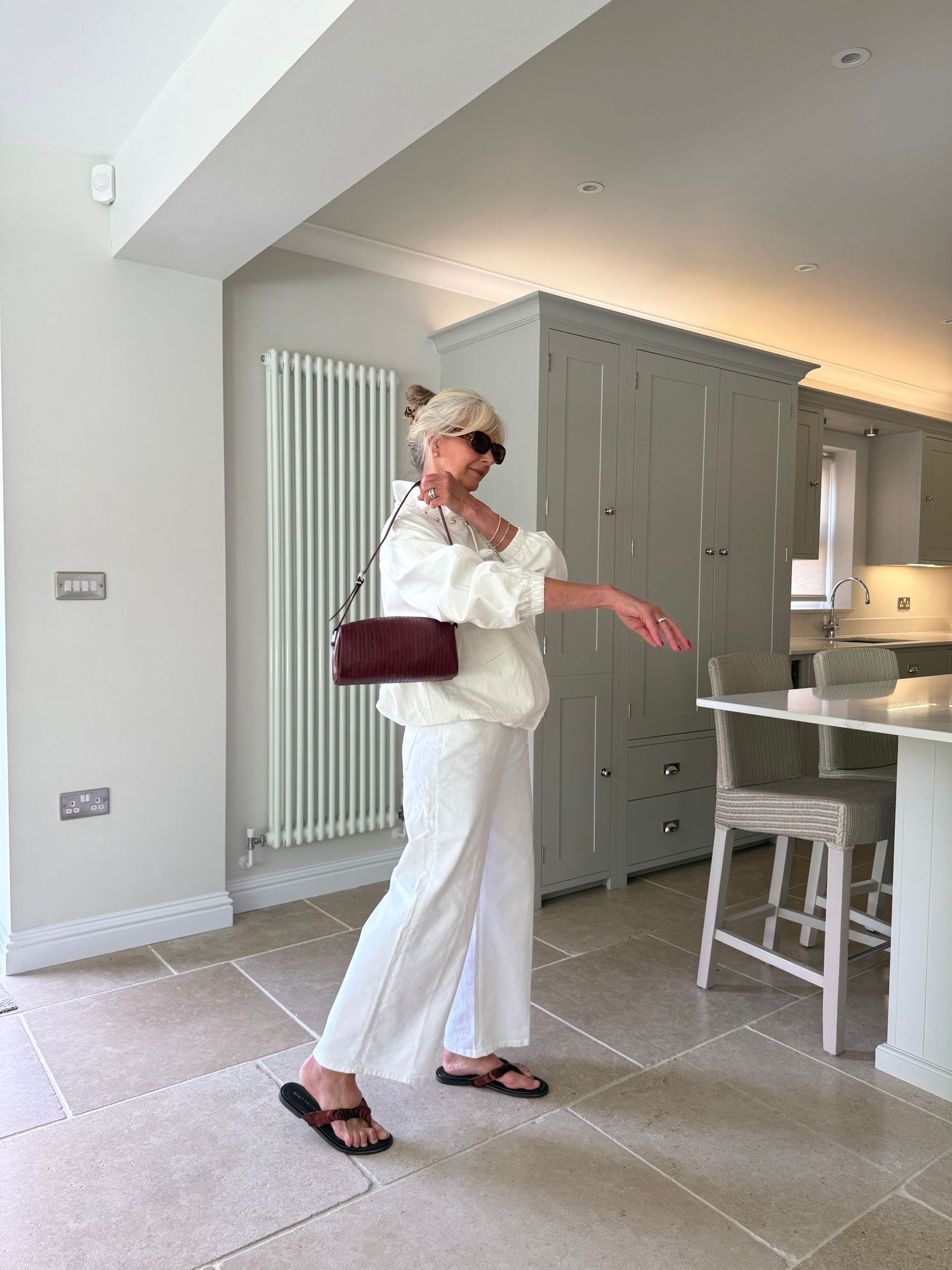 Woman in white linen co-ord set with burgundy shoulder bag and black sandals posing in modern kitchen.