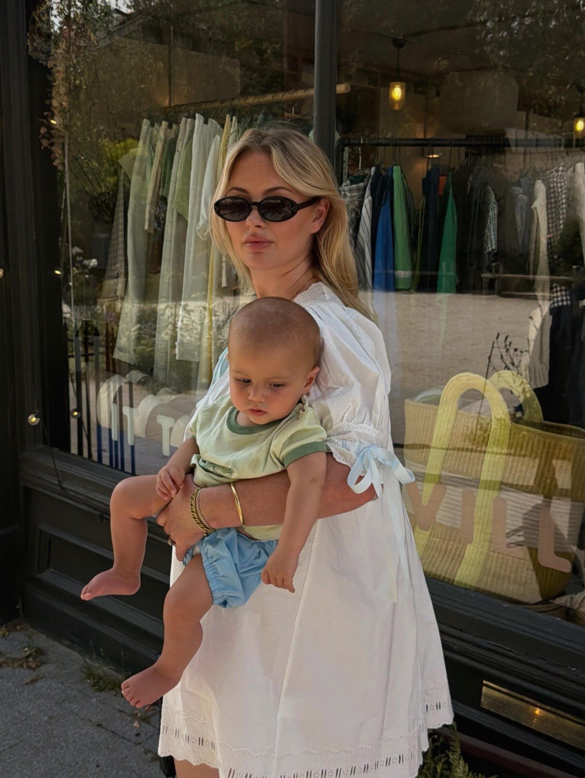 Blonde woman in white dress holding a baby in green outfit, standing outside a boutique store.