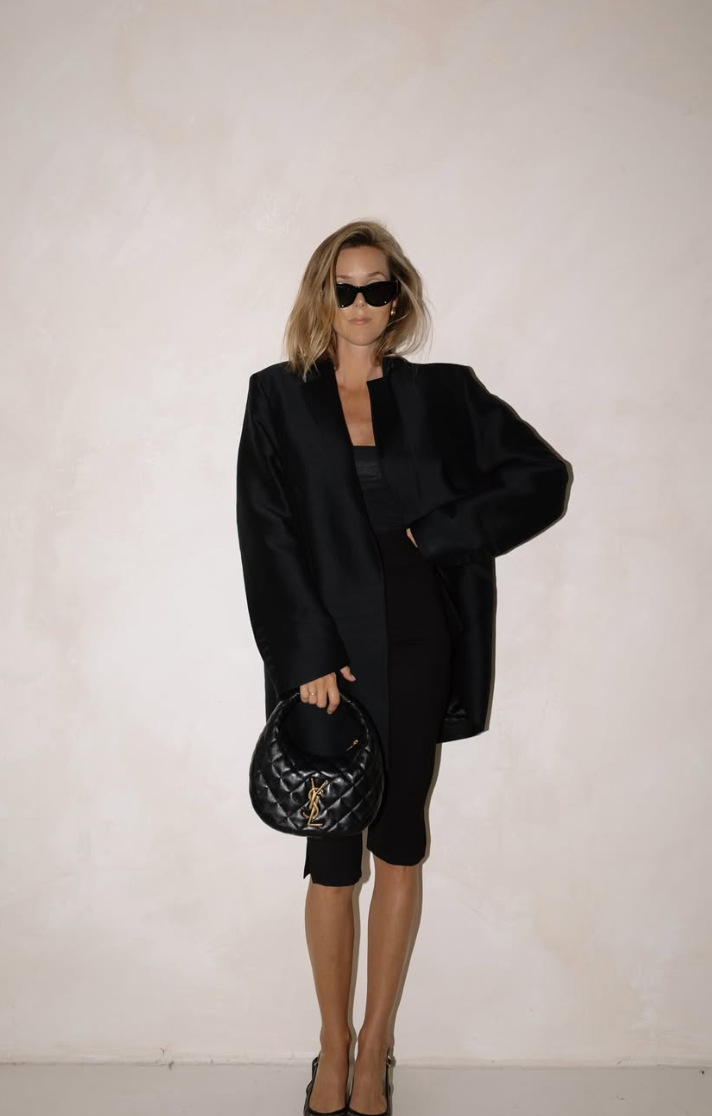 Fashion model in oversized black blazer and shorts poses with designer handbag against white backdrop.