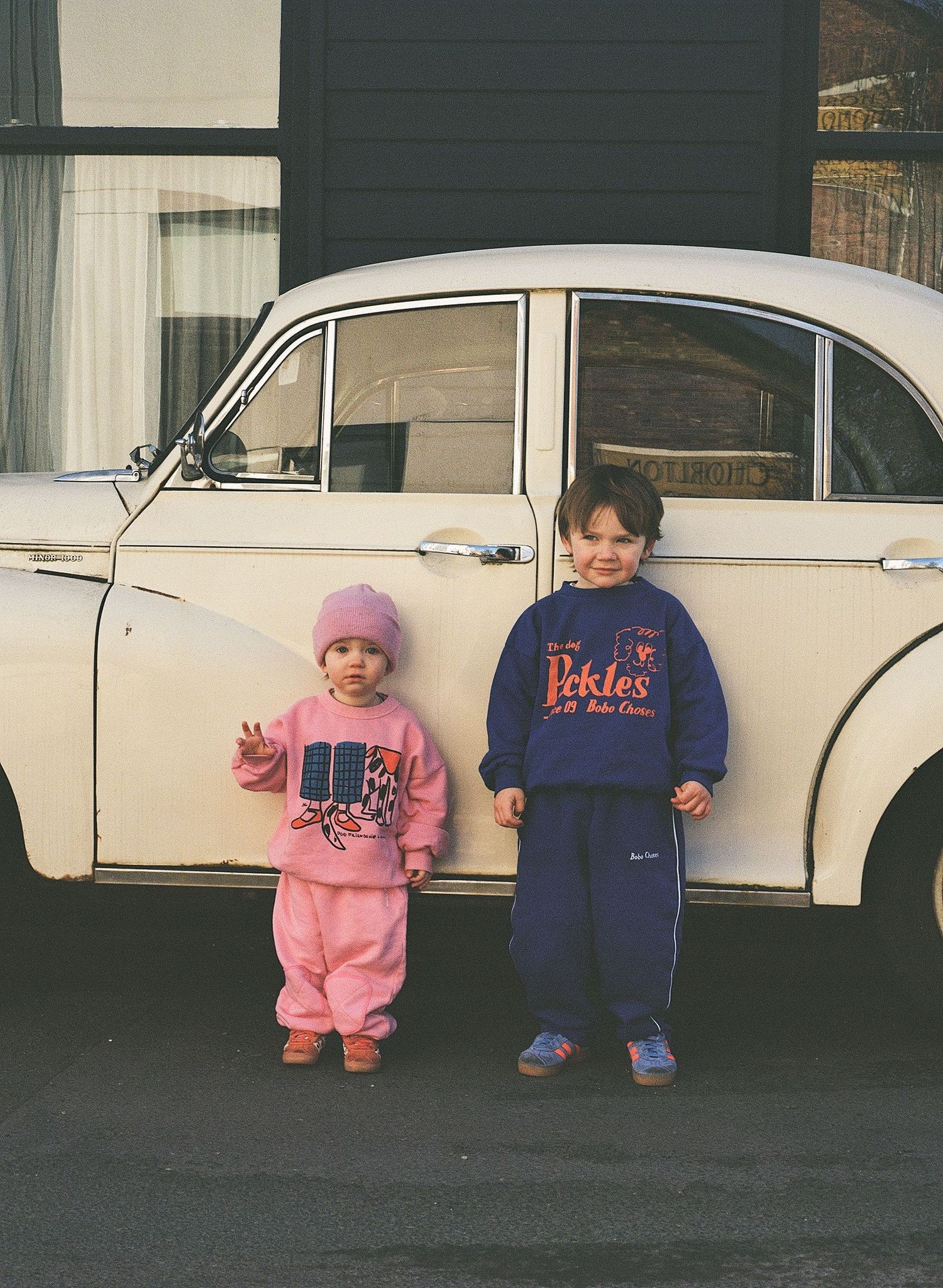 Two small children in pink and blue outfits sit in front of classic white cars in a vintage photograph.