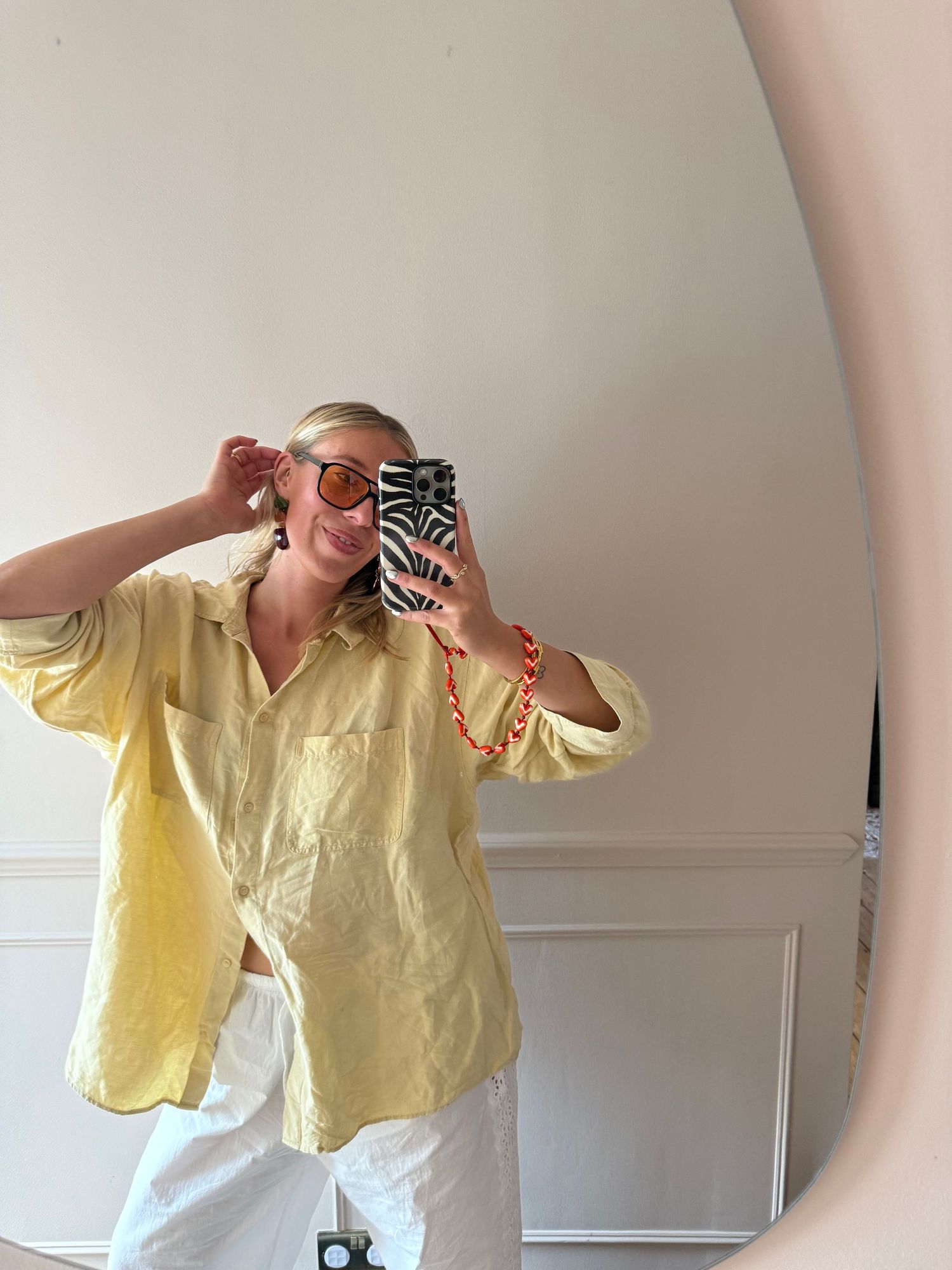 Woman in yellow linen shirt and white pants takes mirror selfie wearing orange sunglasses and red beaded bracelet.