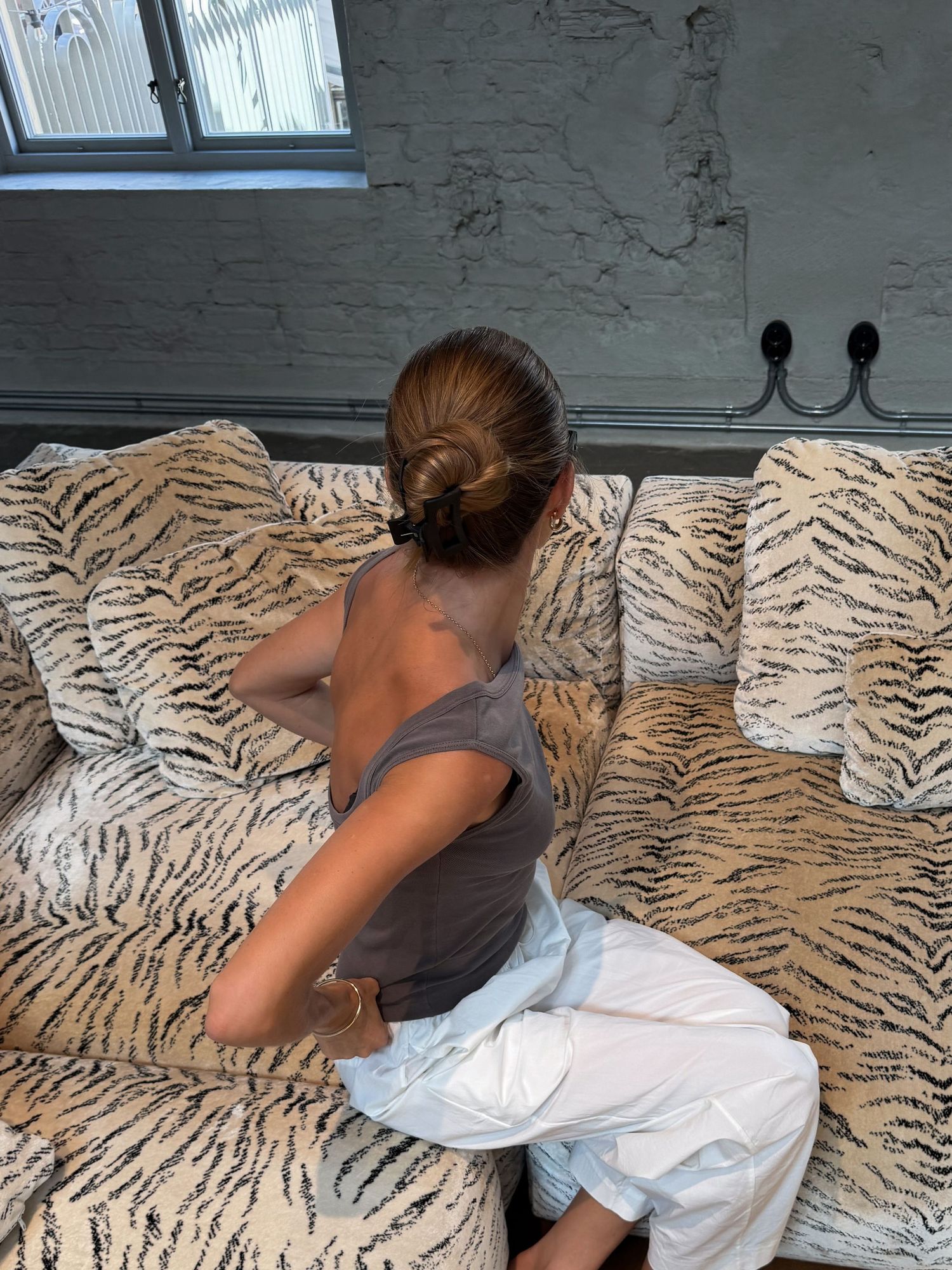 Person with hair bun sitting on a tiger-print sofa, wearing a gray top and white pants.
