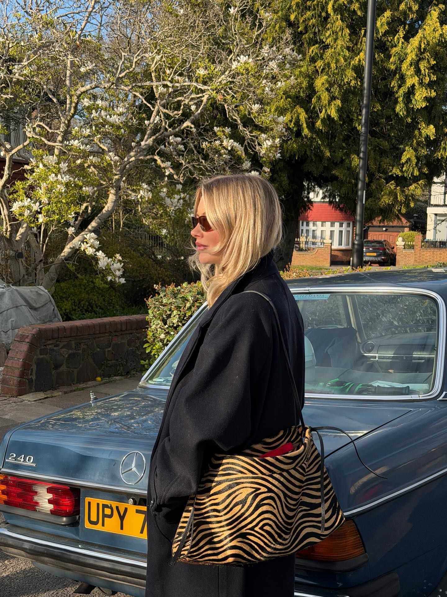 Blonde woman in black coat with zebra print bag beside a vintage Mercedes-Benz on a sunny street.