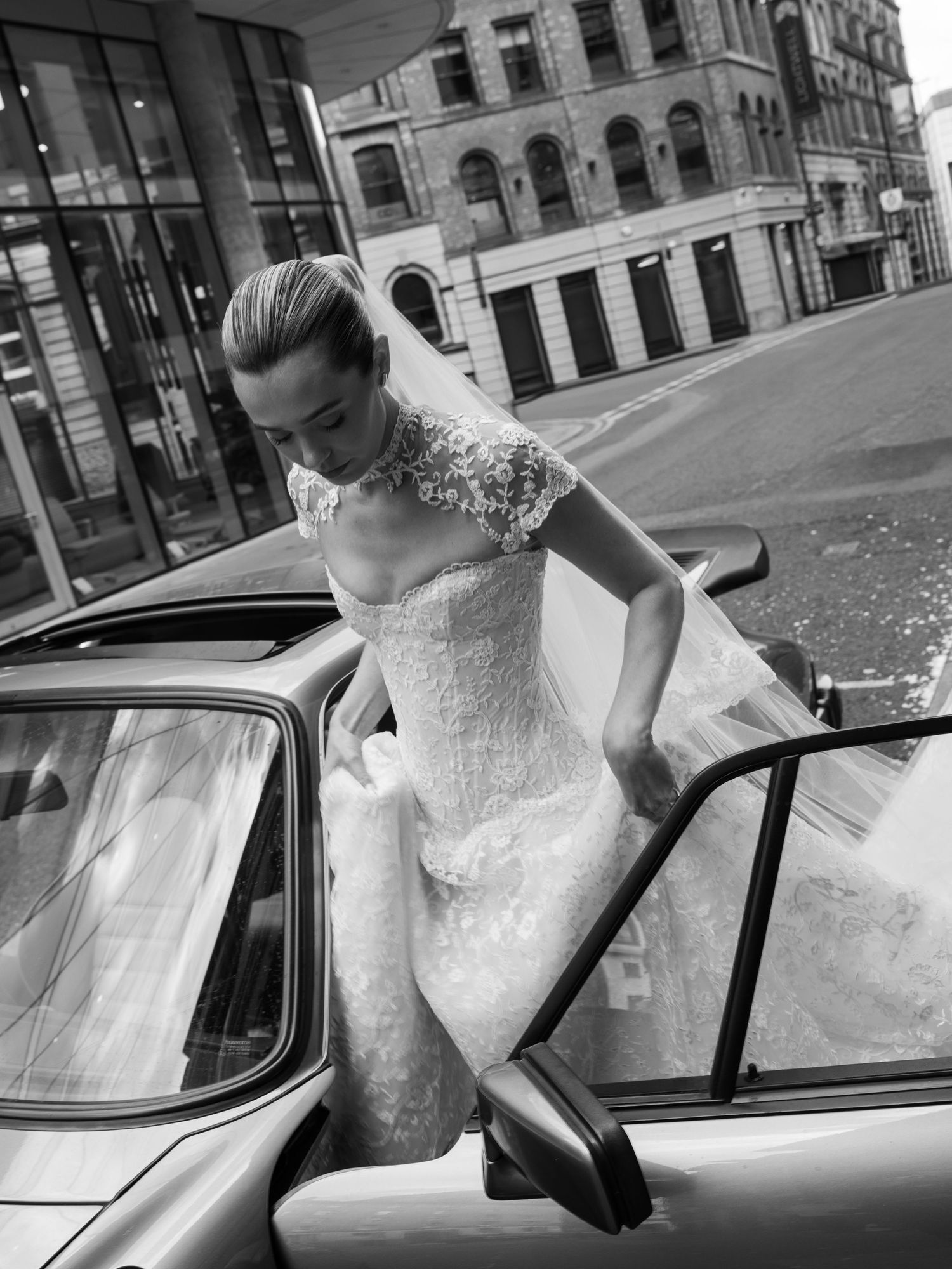 Bride in elegant lace wedding gown stepping out of a classic car on a city street in black and white.