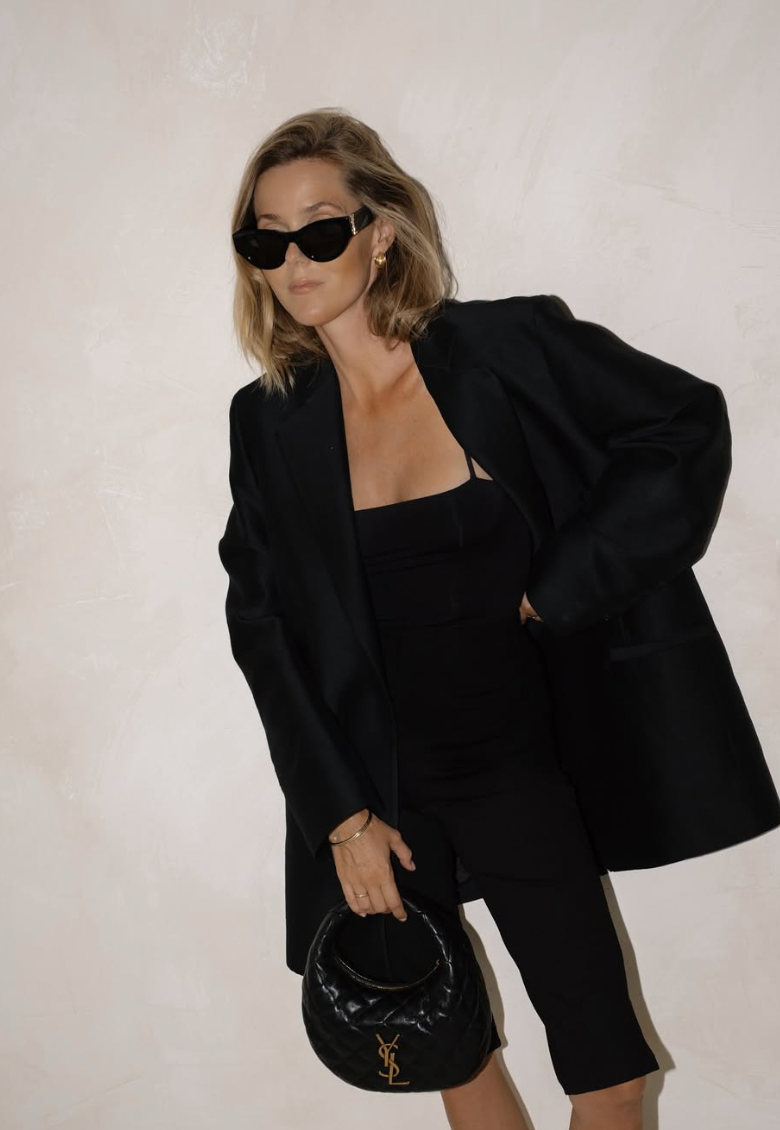 Fashion model in black blazer and sunglasses poses against white background in stylish editorial series.