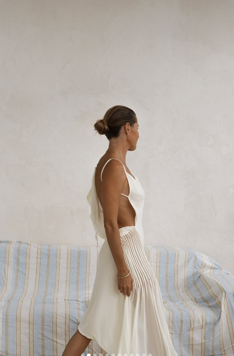 Woman in white dress poses against textured wall with striped fabric backdrop in minimalist studio setting.