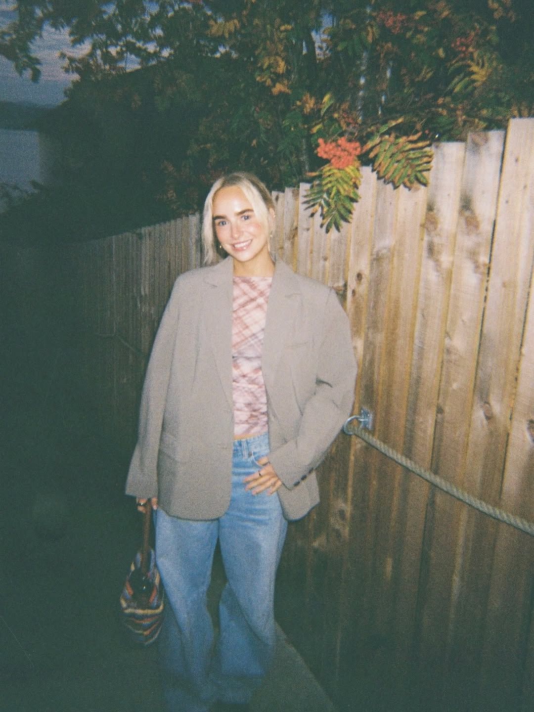 Blonde woman smiling in oversized blazer, pink top, and blue jeans, standing by a wooden fence outdoors.
