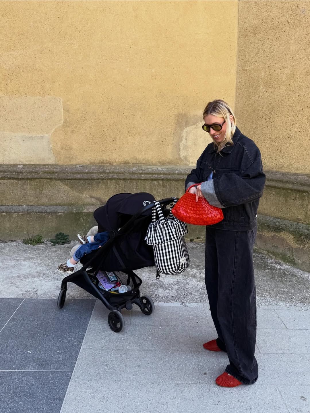 Woman in black jumpsuit with red bag and shoes pushes stroller on urban sidewalk near yellow wall.