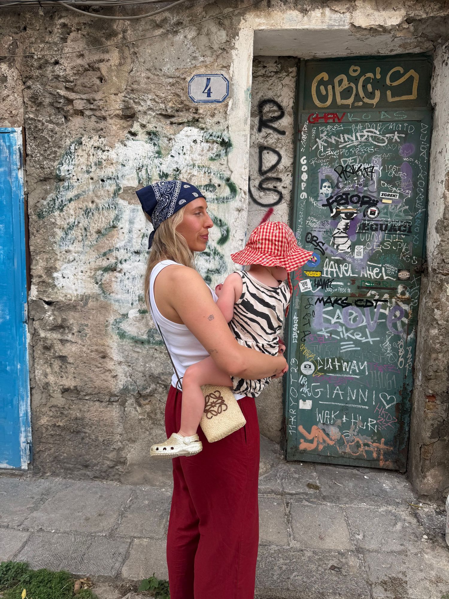 Woman holding a baby in a gingham hat near a graffiti-covered door on a rustic European street.
