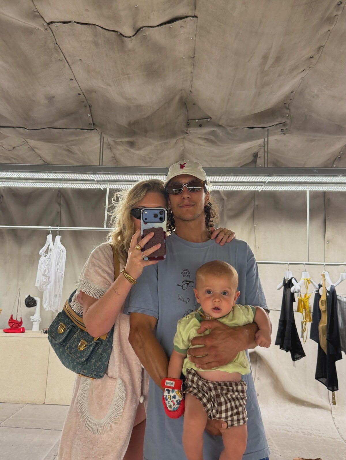 Young couple taking mirror selfie with baby in arms, standing under tent with clothing rack in background.