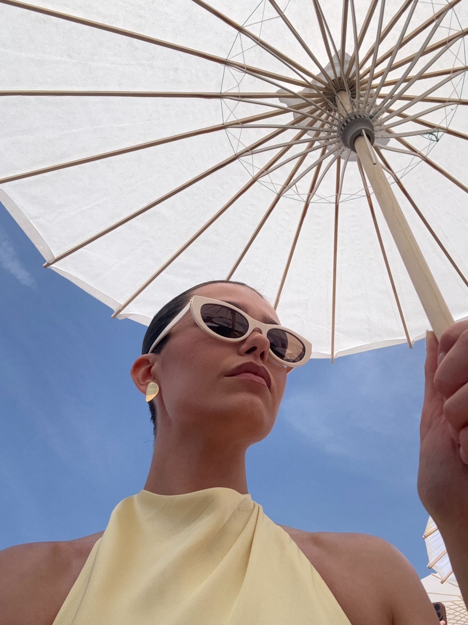 Woman in yellow halter top and white sunglasses relaxing under a white beach umbrella against a blue sky.