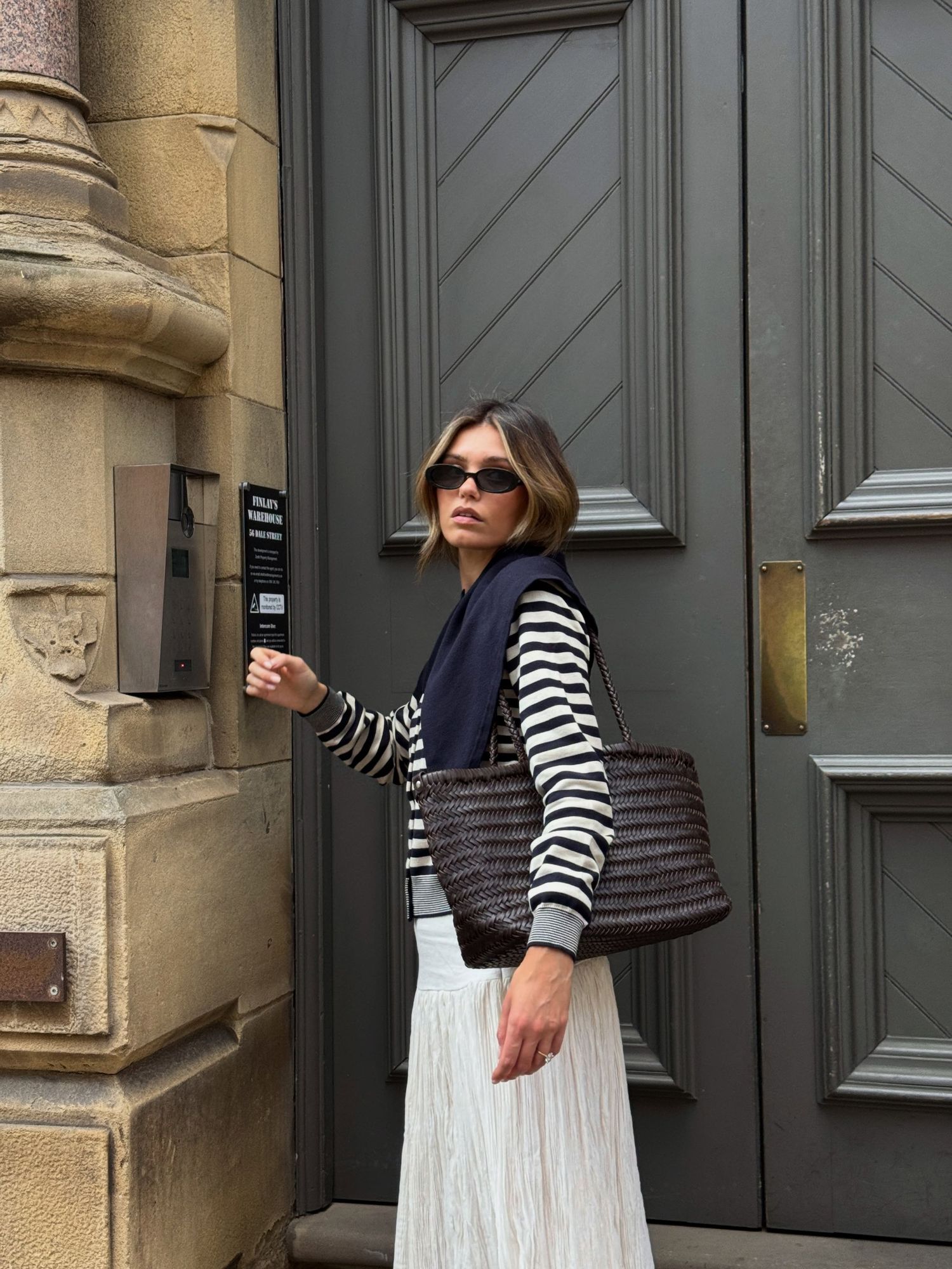 Woman wearing striped shirt, navy vest, white pleated skirt, carrying a brown woven tote bag outdoors.
