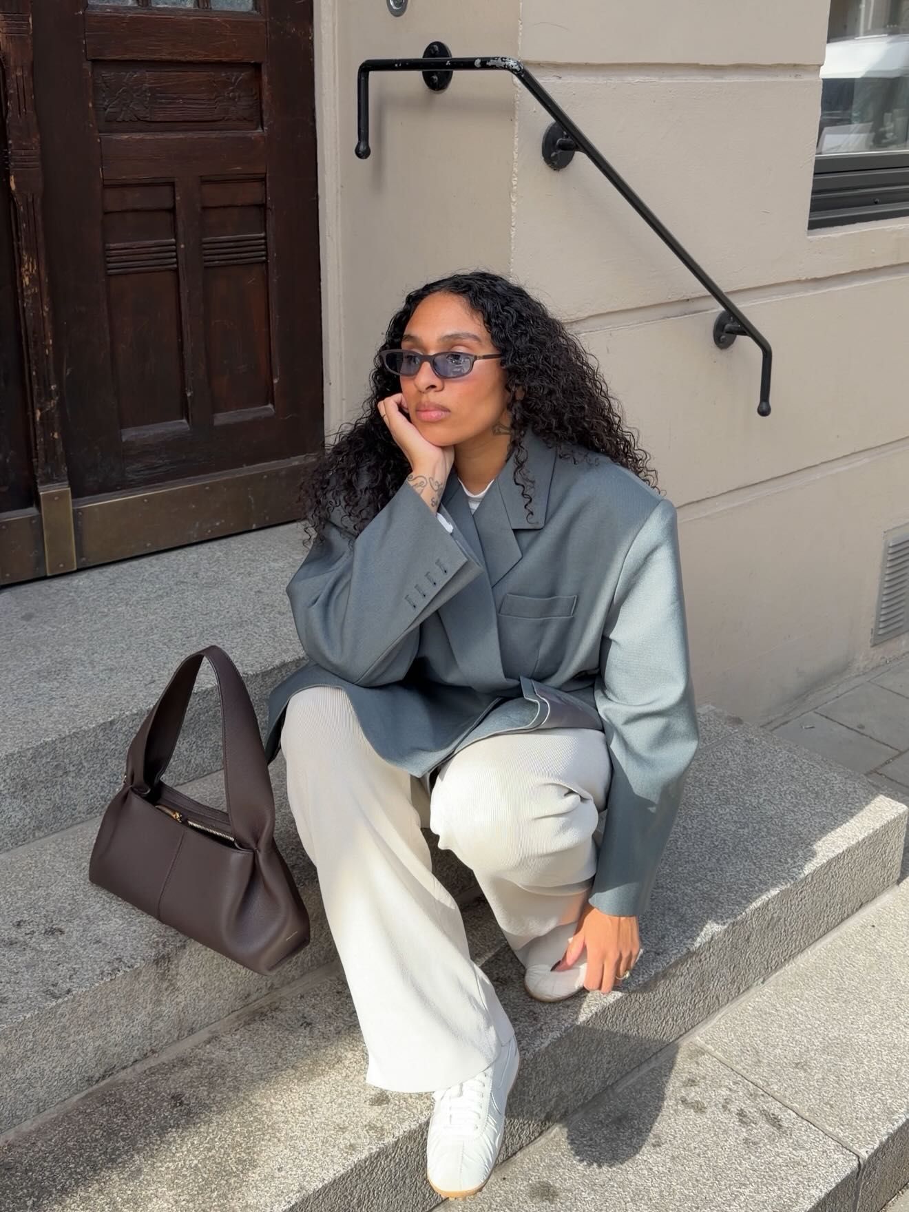 Woman in gray blazer, cream wide-leg pants, and sunglasses sits on steps with a brown leather bag.