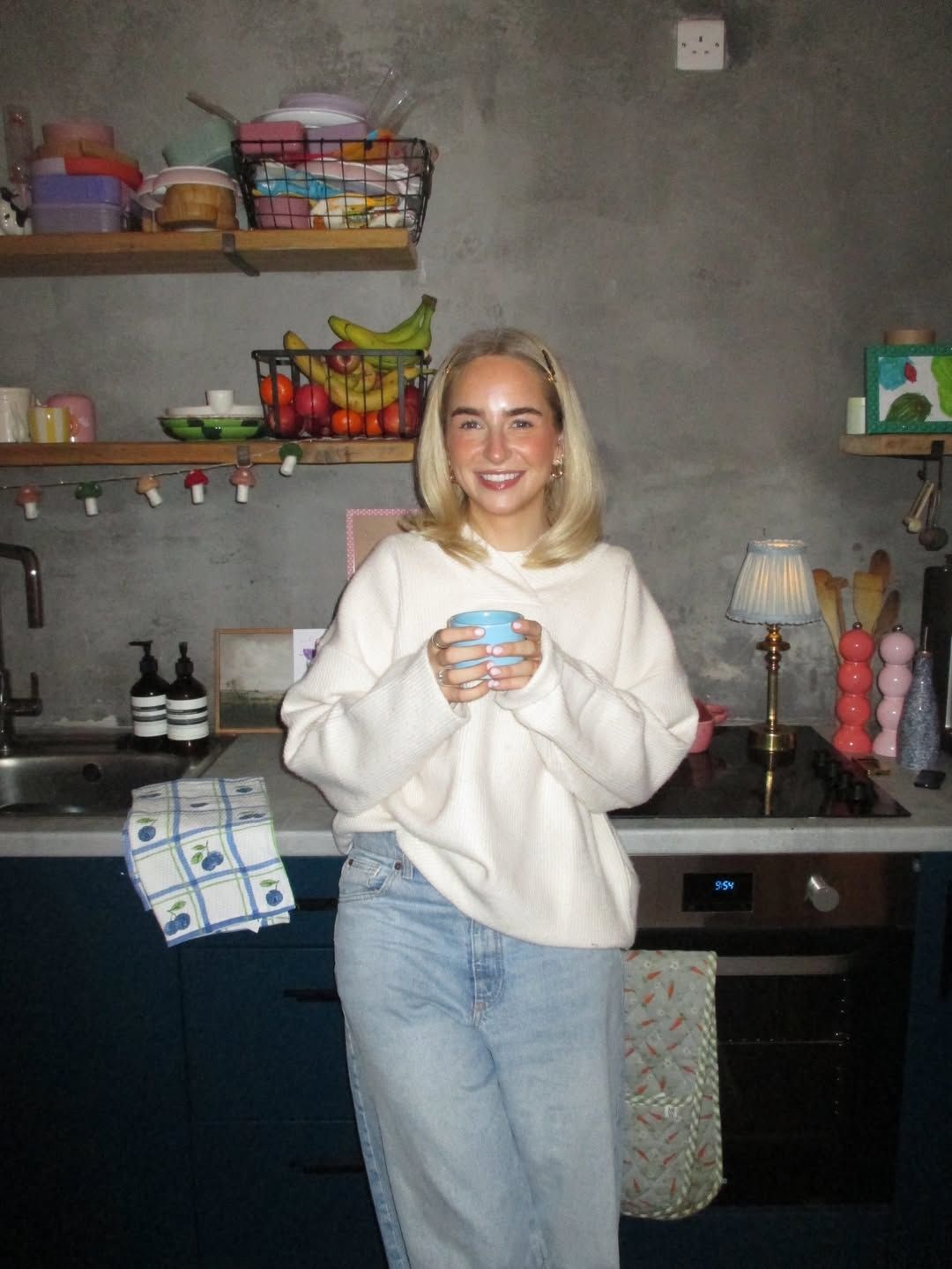 Smiling blonde woman in cream sweatshirt holding blue mug in stylish dark kitchen with colorful shelves.